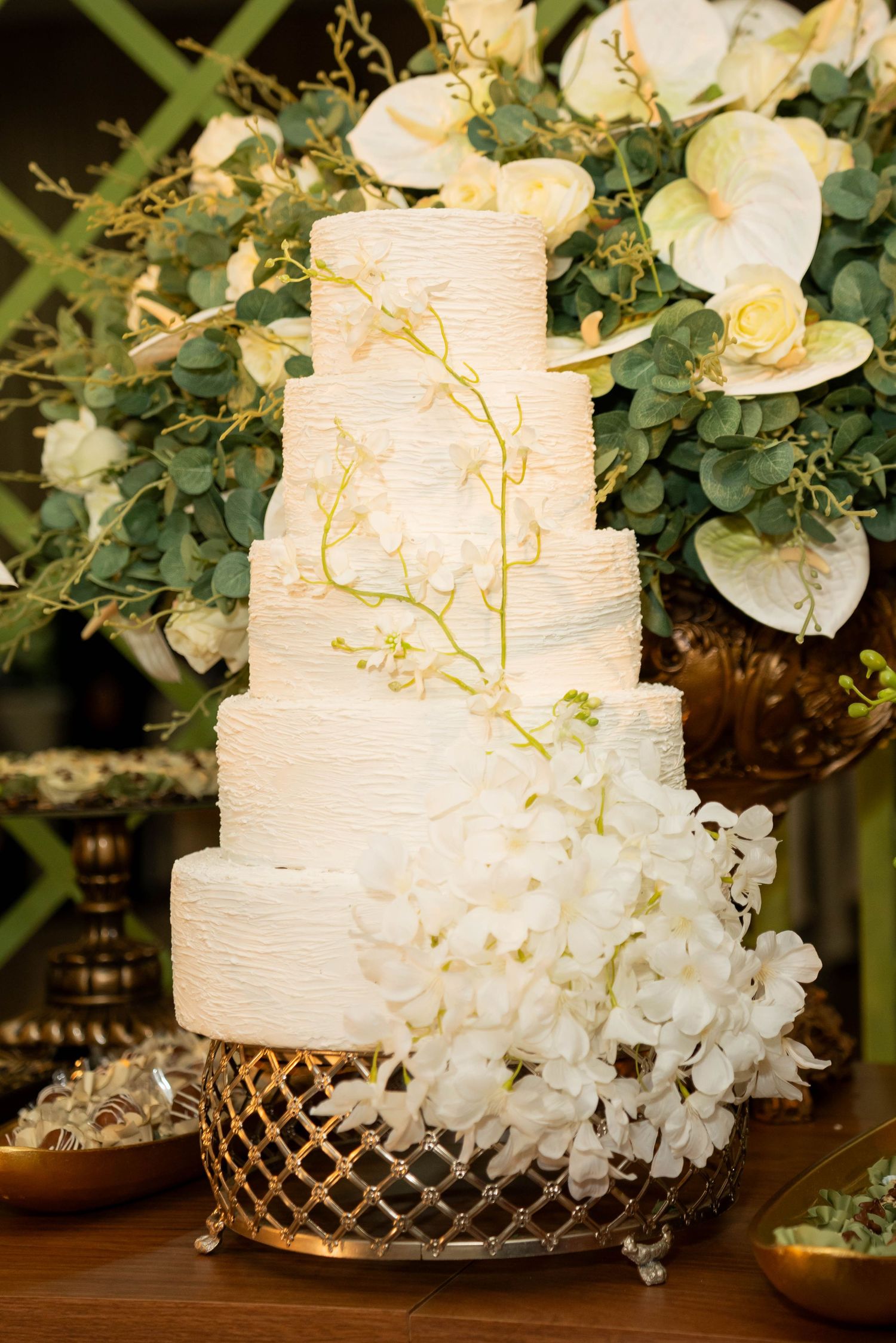 Elegant four-tier white wedding cake with textured frosting, white orchids, and gold stand, displayed against lush floral backdrop.
