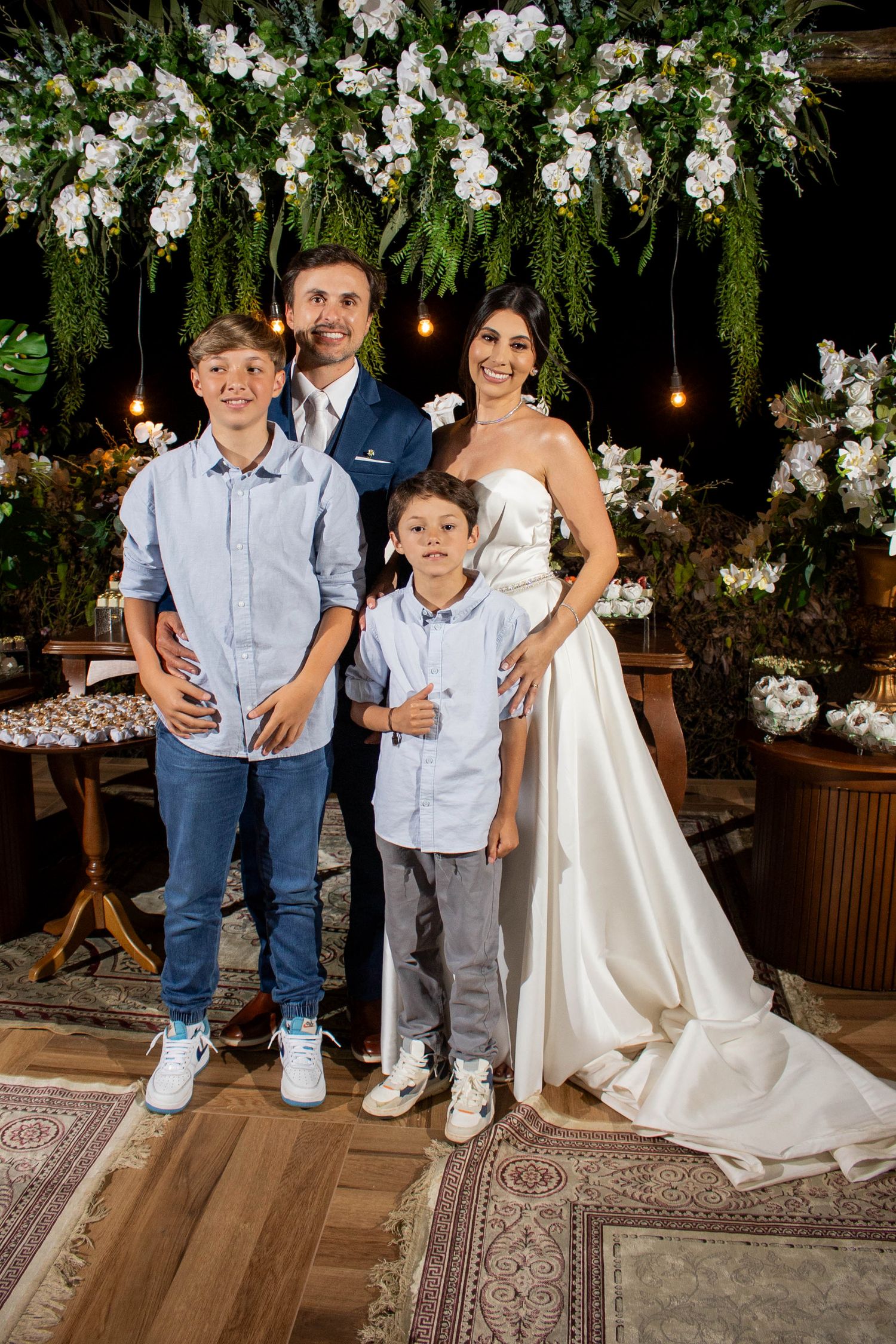 A family of four poses at a wedding, with the bride in white, groom in navy, and two boys in blue shirts.