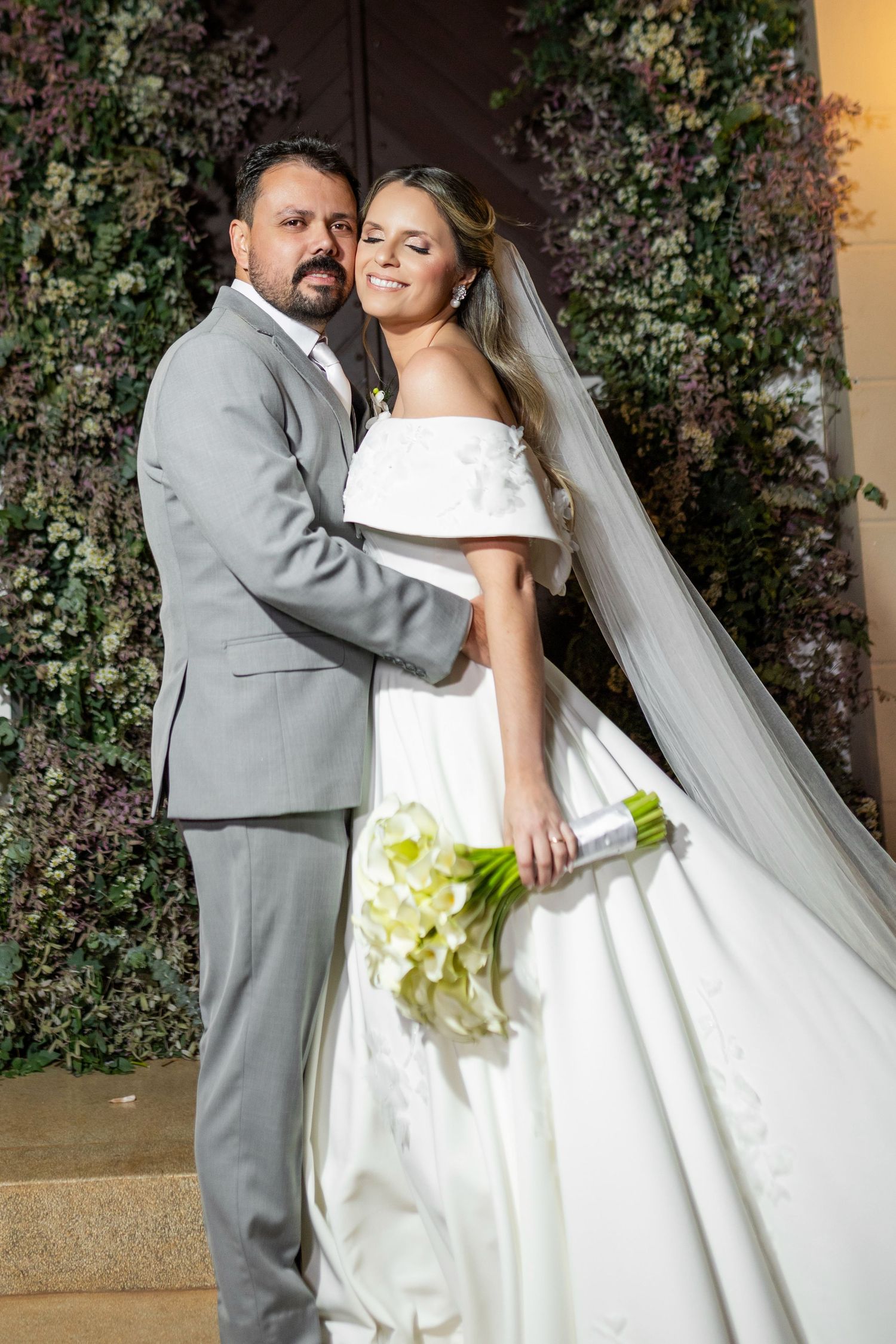 A bride and groom pose elegantly before a lush green floral wall, she holds white calla lilies wearing an off-shoulder gown.