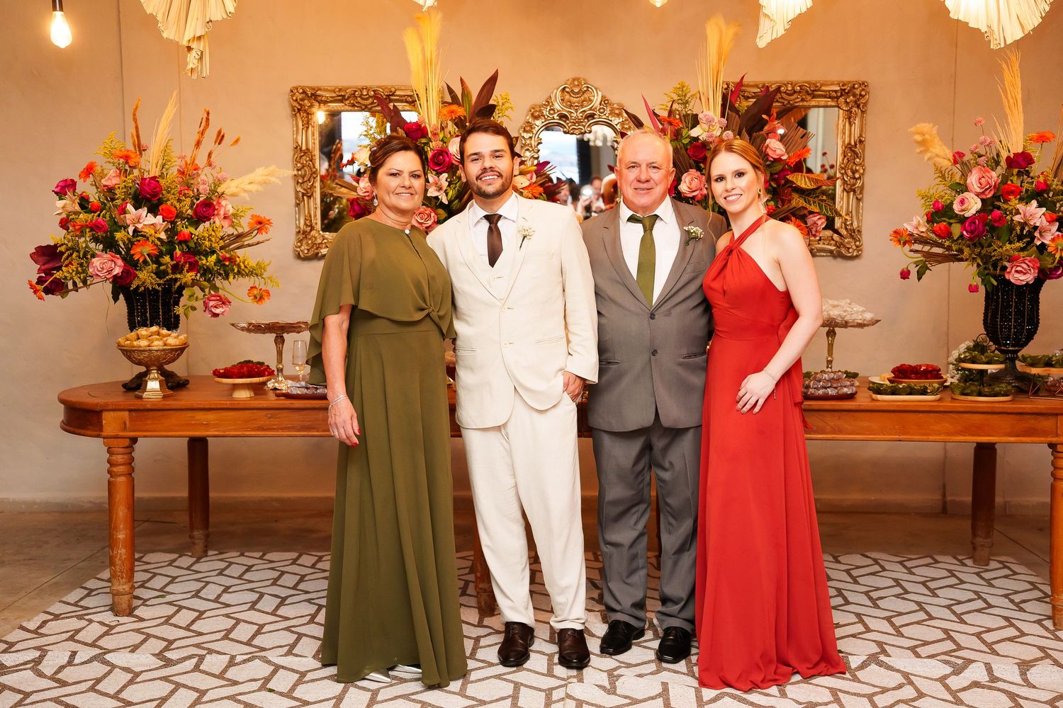 Four elegantly dressed people posing together at a formal event with floral arrangements and ornate mirrors in background.