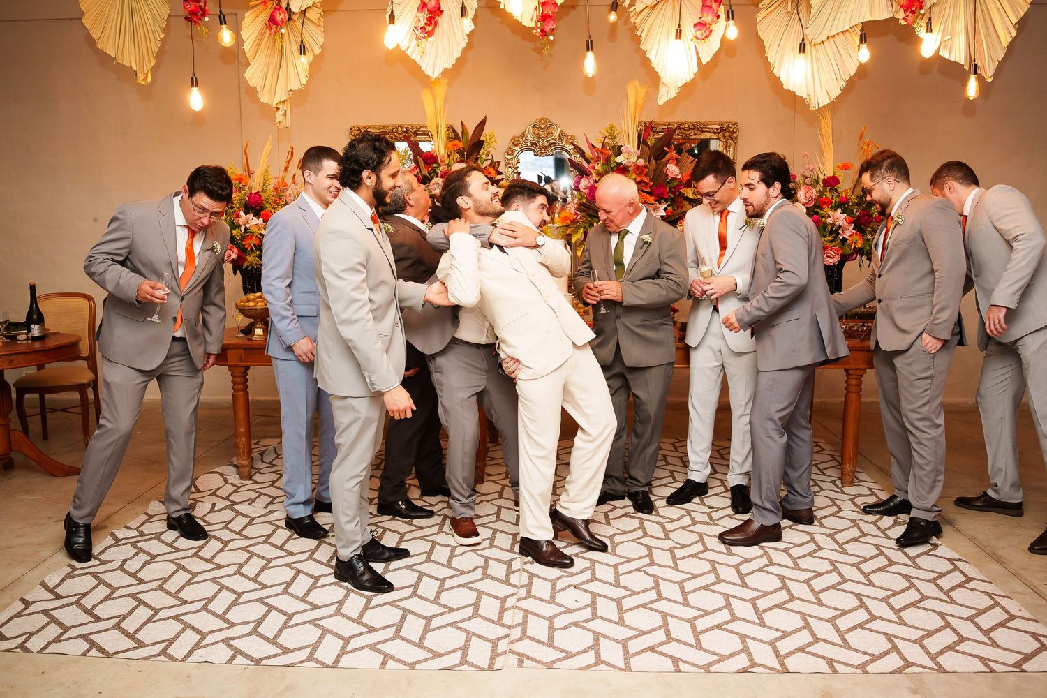 Groomsmen in gray suits celebrate with the groom in a white suit, laughing and dancing at a festive wedding reception.