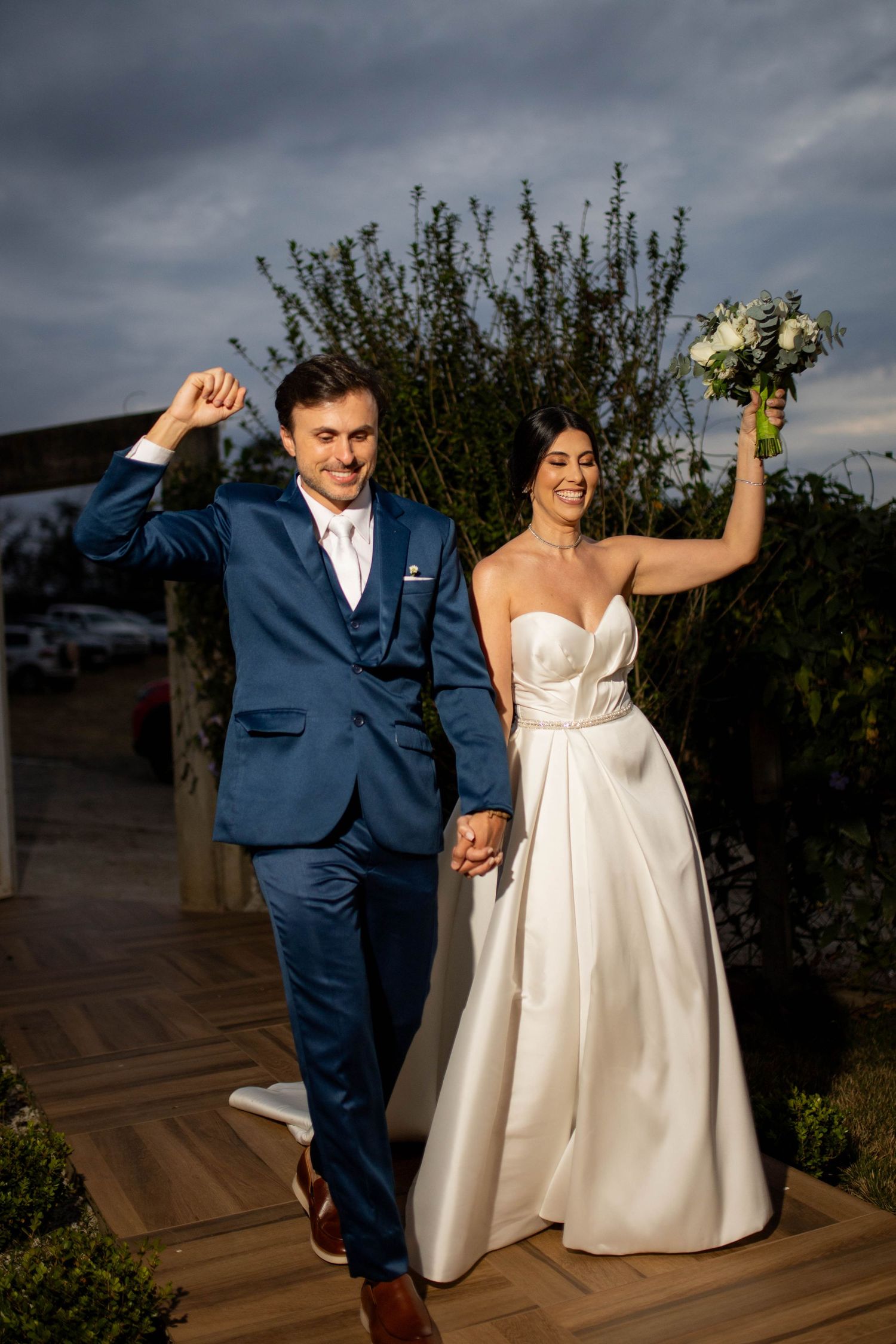 Joyful newlyweds celebrate after wedding ceremony, groom in navy suit, bride in strapless white gown holding bouquet.