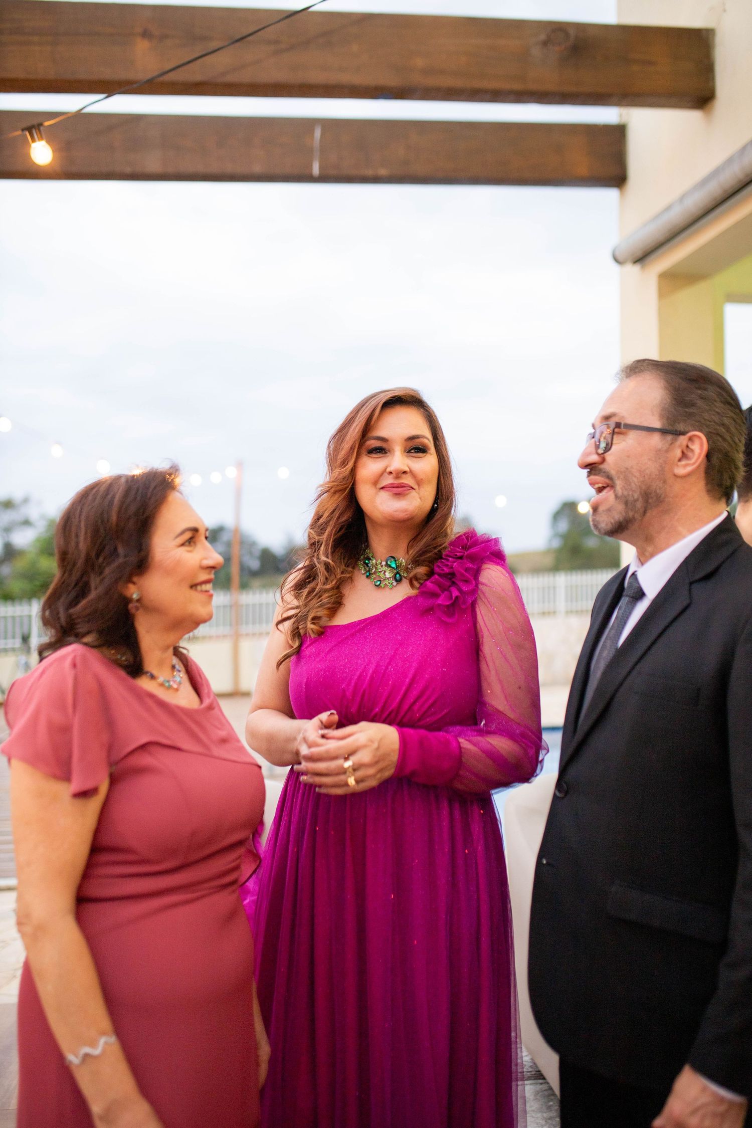 Three elegantly dressed people socializing at a formal event, women in pink and magenta gowns, man in black suit.