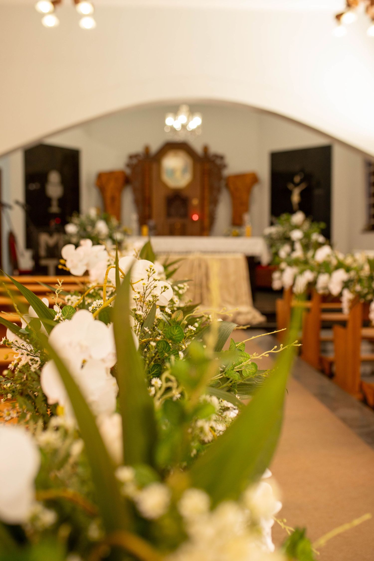 Church aisle decorated with white flowers and greenery leading to ornate wooden altar for a wedding ceremony.