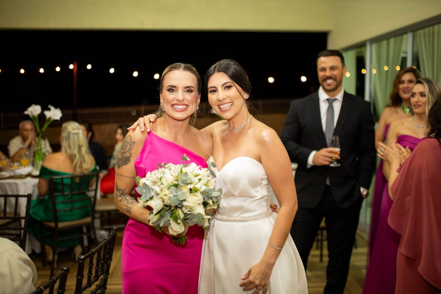 Bride in white strapless gown poses with guest in pink dress holding bouquet at elegant wedding reception.