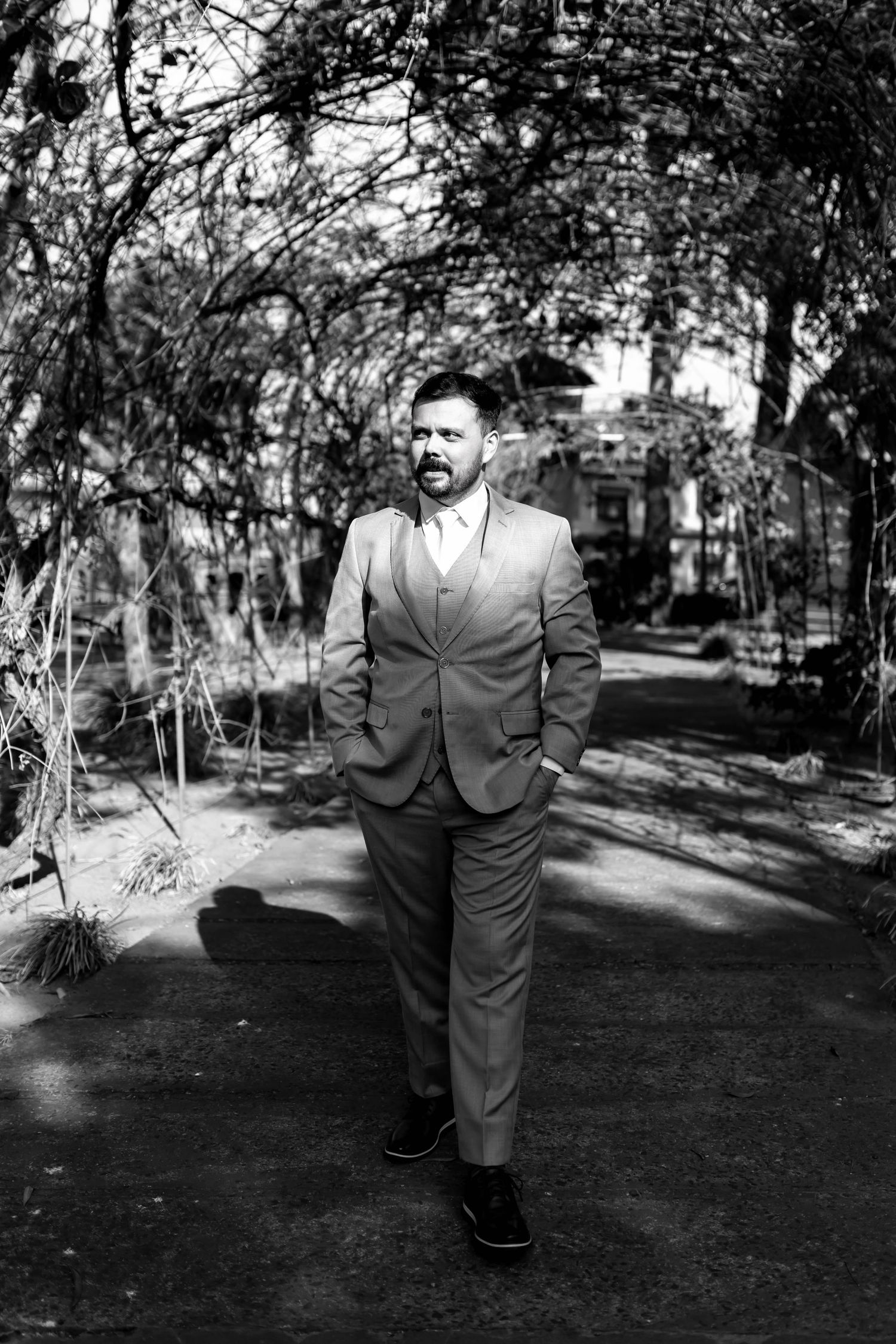 Black and white portrait of a bearded man in a tailored suit walking confidently through a garden pathway.