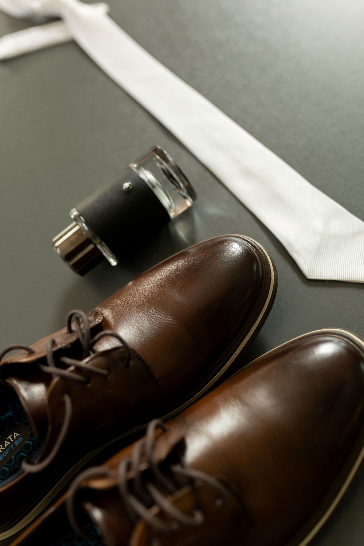 Brown leather dress shoes, white tie, and cologne arranged on dark surface for a formal mens outfit flatlay.