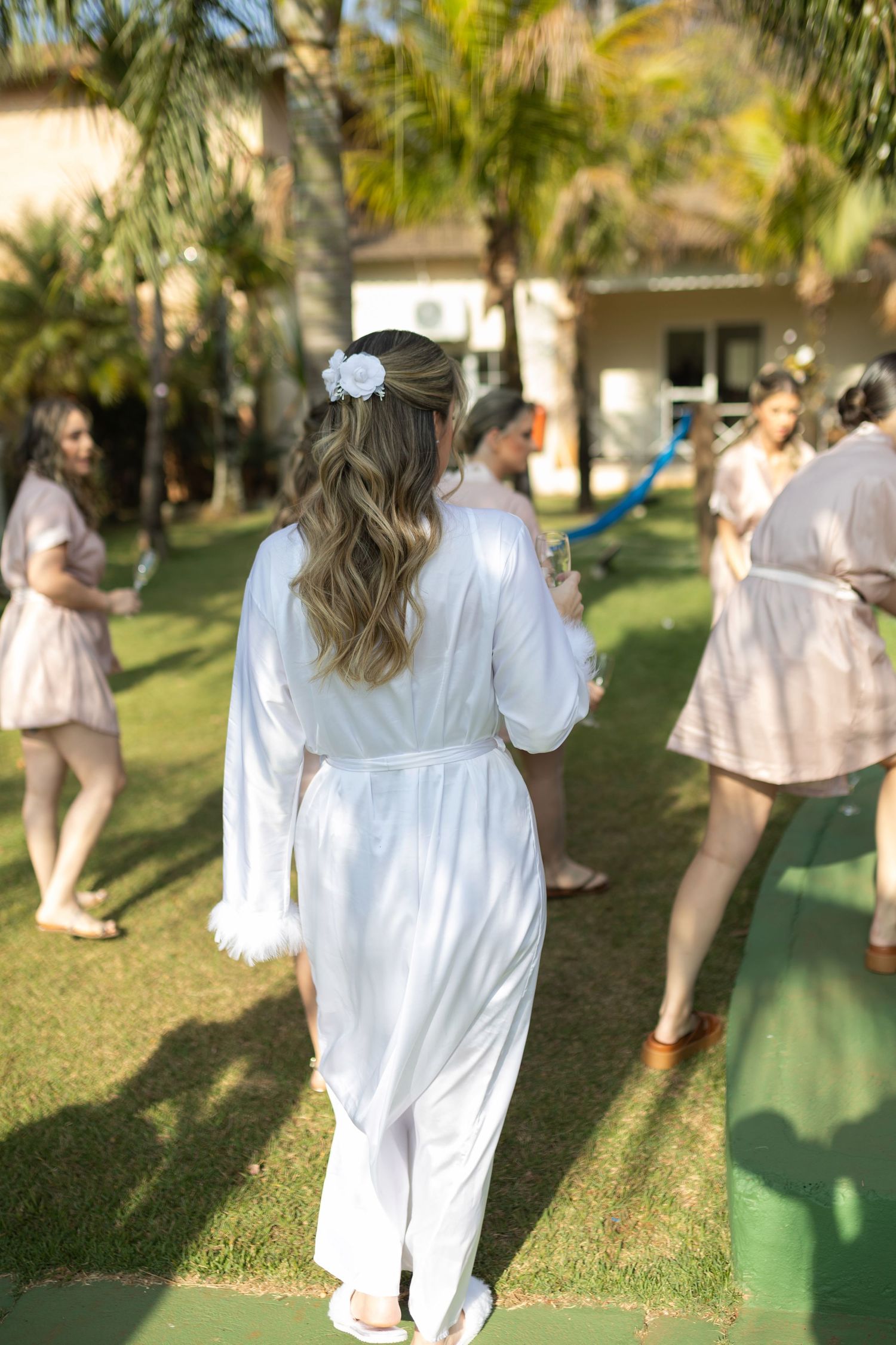 Bride in white robe with flower hair accessory stands on green lawn with bridesmaids in pink robes at outdoor pre-wedding celebration.