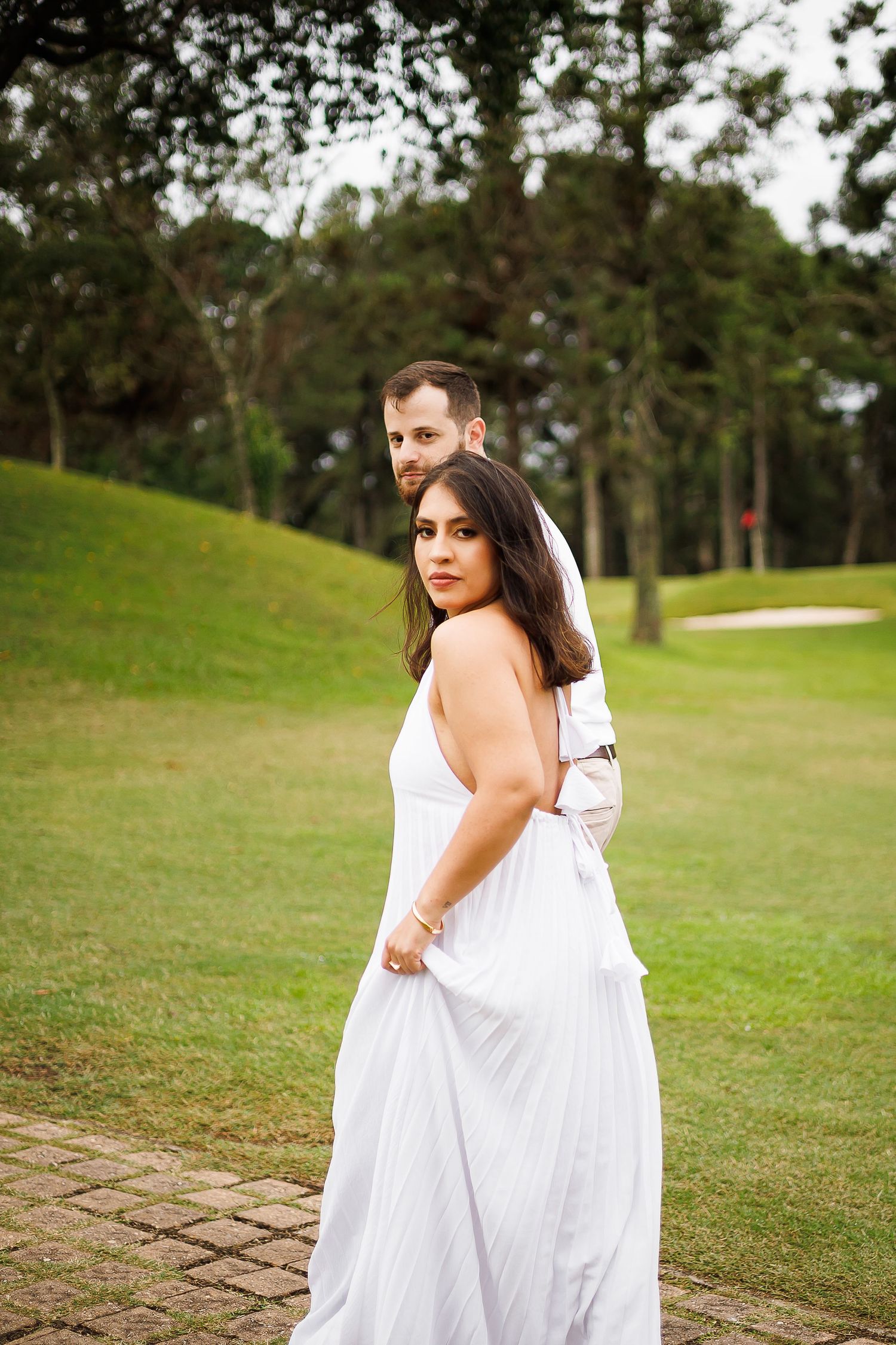 Couple in white attire posing on lush green golf course, woman in elegant white gown looking back at camera.
