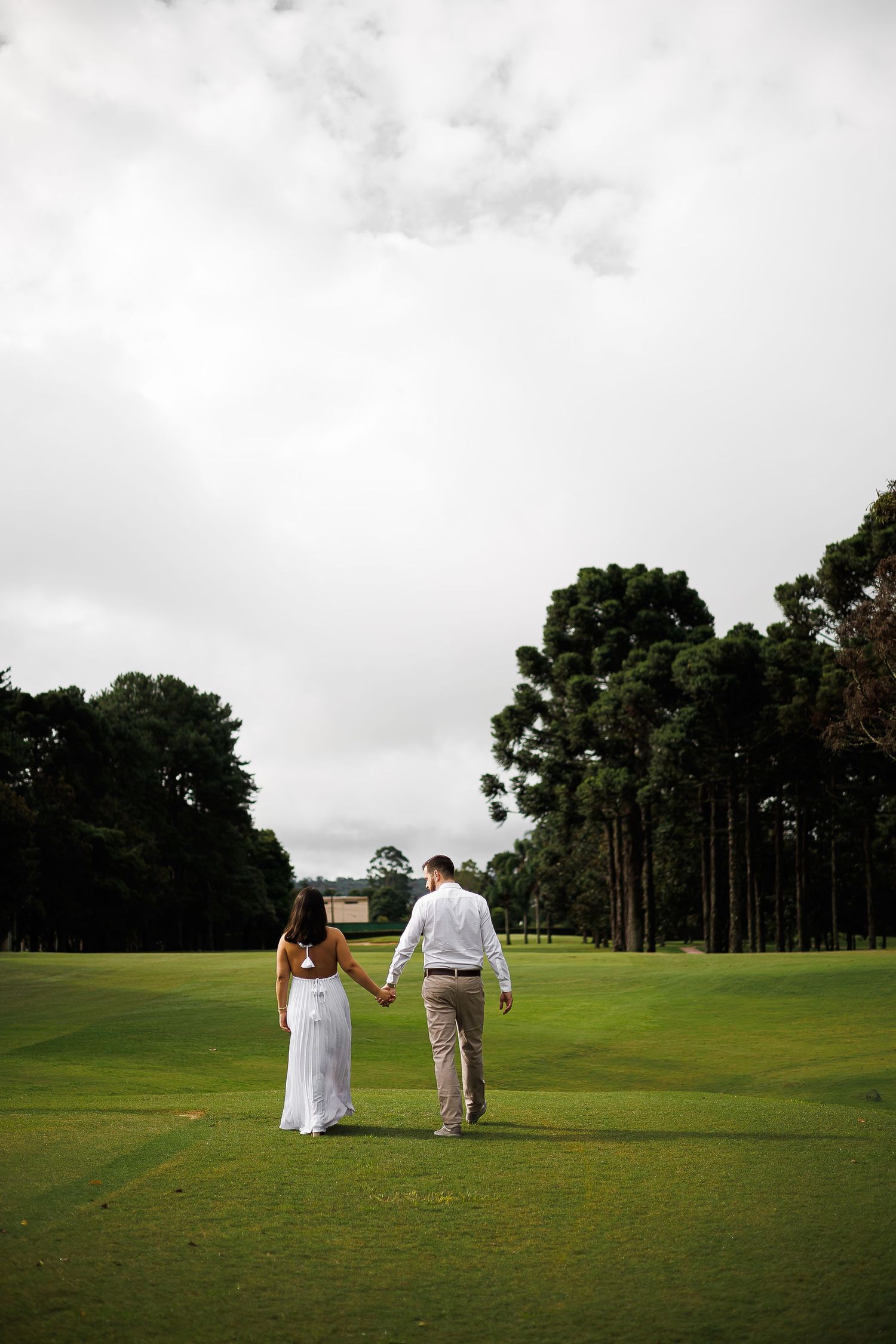 Couple in wedding attire holding hands while walking on a lush green golf course surrounded by tall trees.