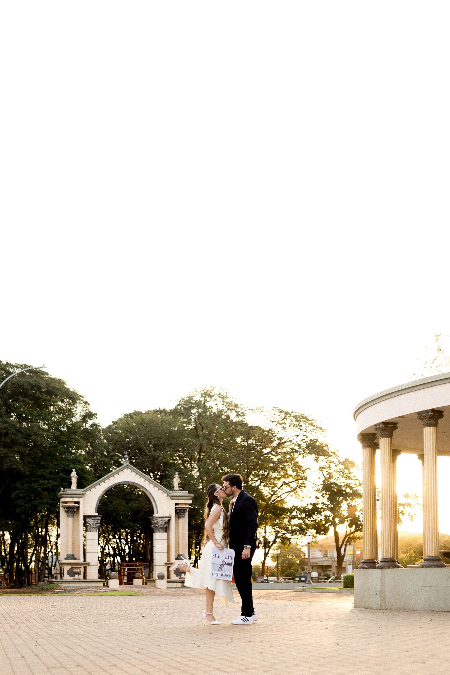 Couple sharing a romantic moment in a sunlit plaza with classical arch and colonnade architecture surrounded by trees.