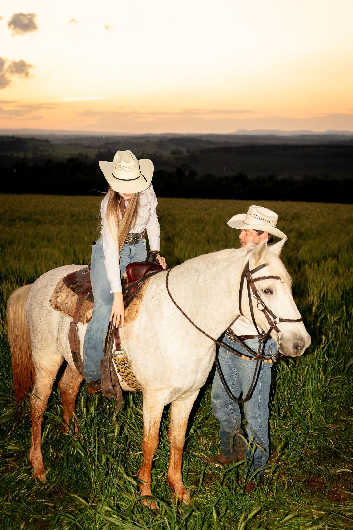 Two cowgirls in white hats, one riding a white horse in a green field at golden sunset.