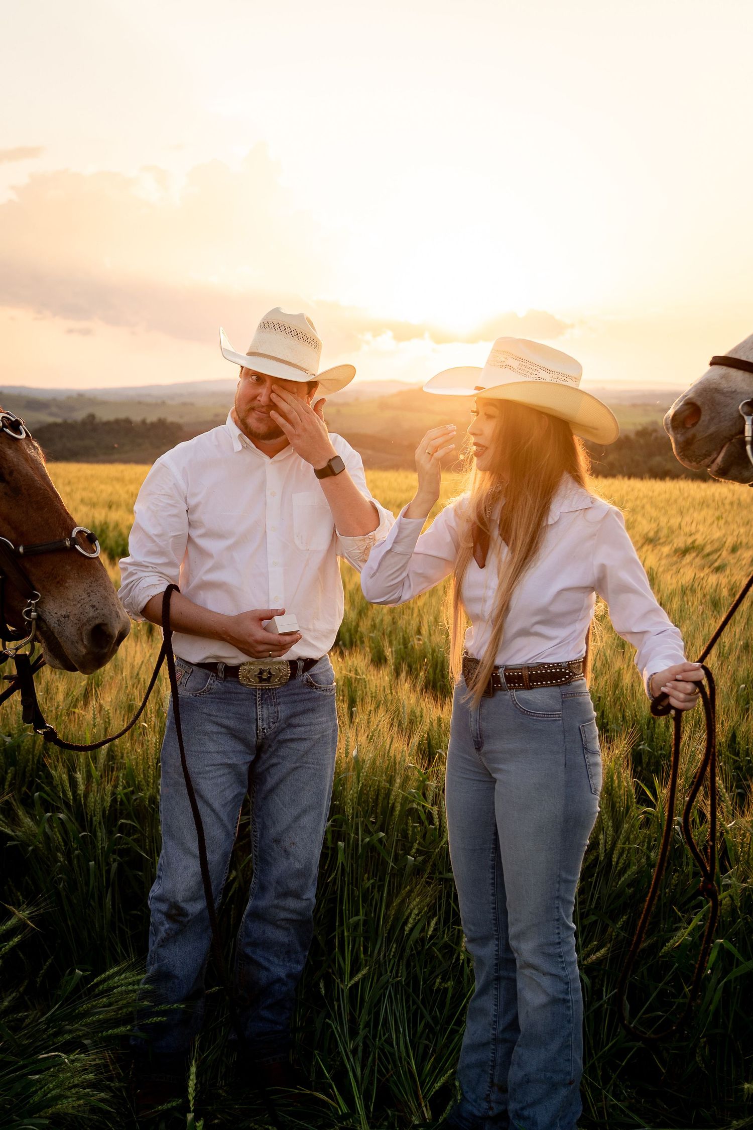 A couple in white shirts, cowboy hats, and jeans stand with horses in a golden sunset field.