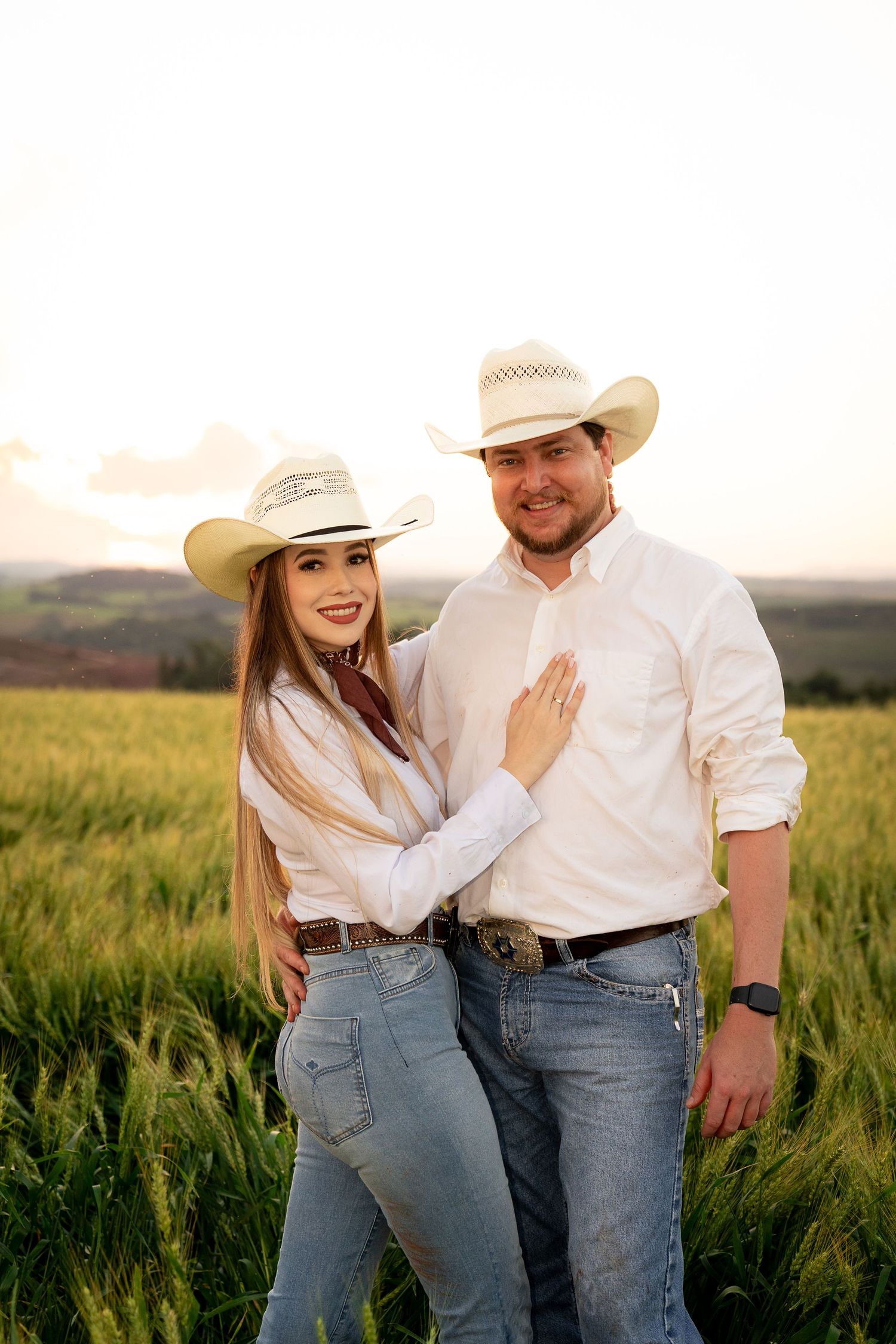 A couple in white cowboy hats and jeans pose together in a green field at sunset.