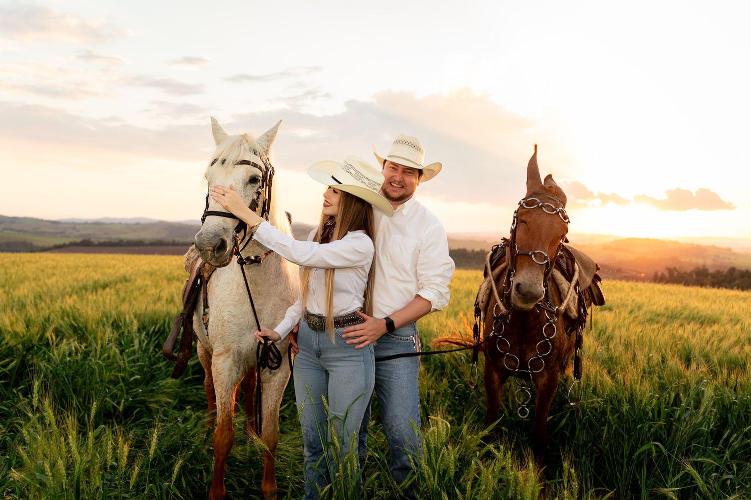 Couple in cowboy hats standing between two horses in a green field at sunset.