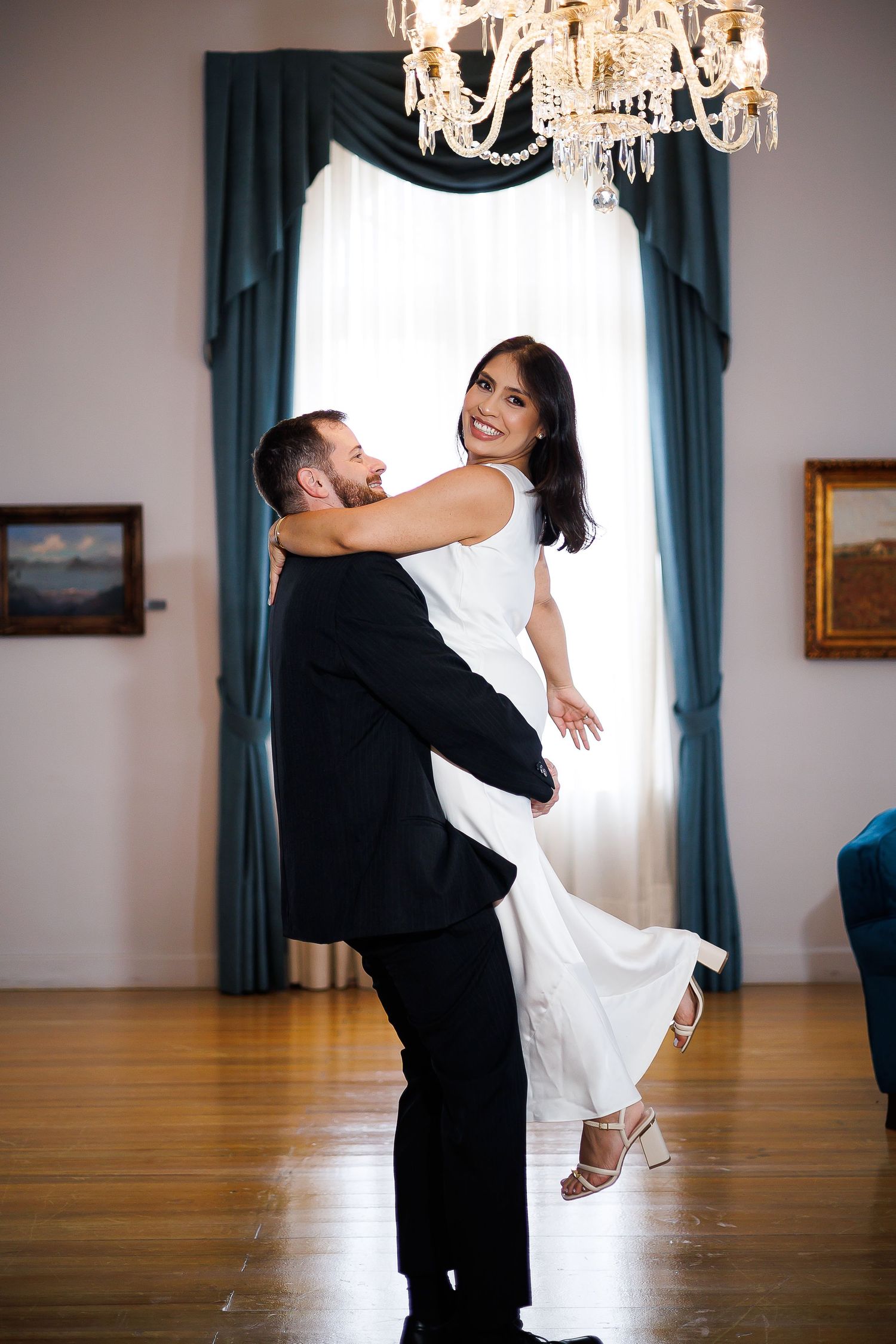 Groom lifting bride in white dress, laughing joyfully in elegant ballroom with chandelier and teal drapes.