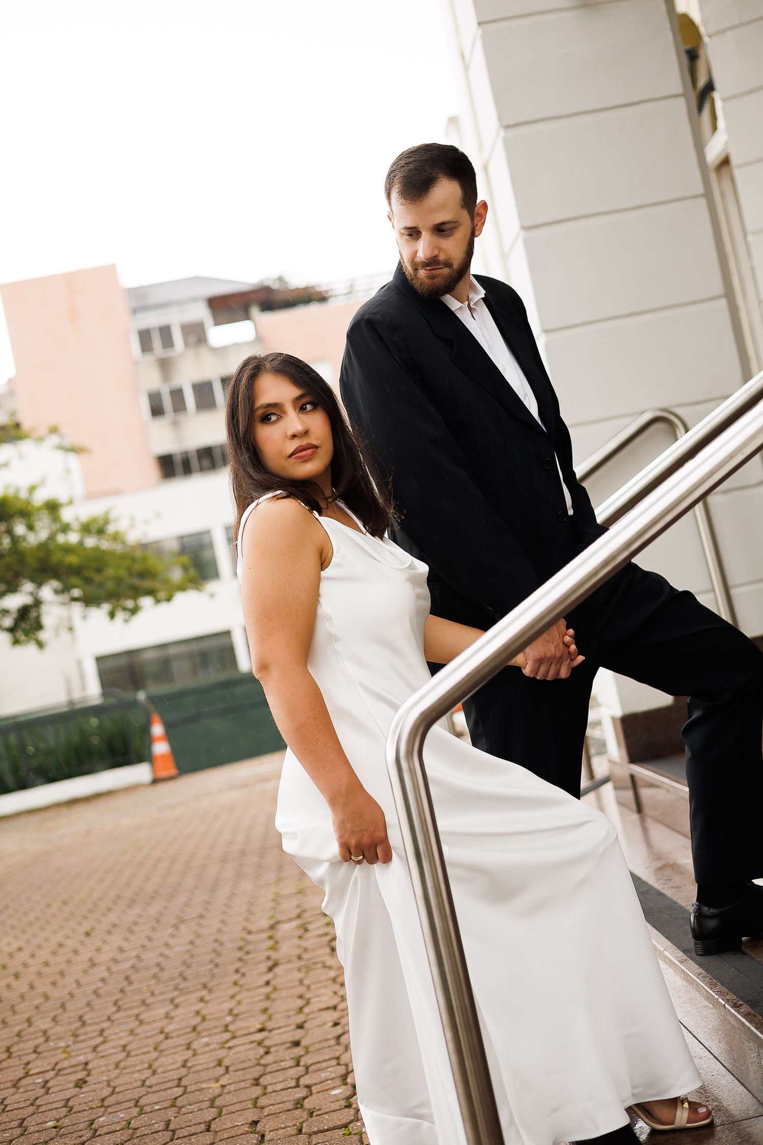 Elegant couple in formal attire posing on outdoor stairs, woman in white gown and man in black suit holding hands.
