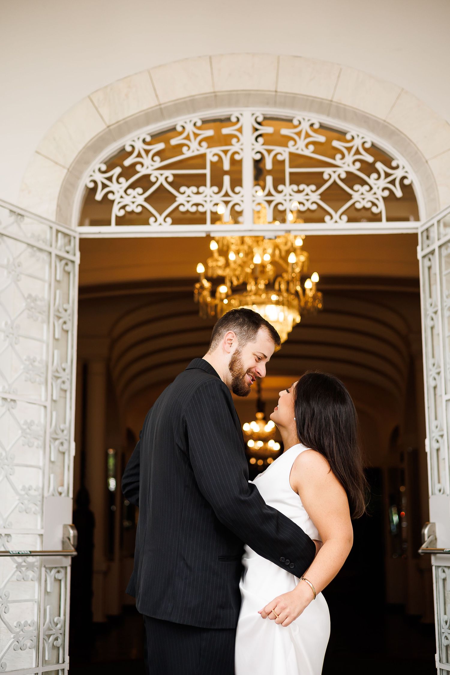 Couple embracing in front of ornate white arched doorway with chandelier, man in black suit, woman in white dress.