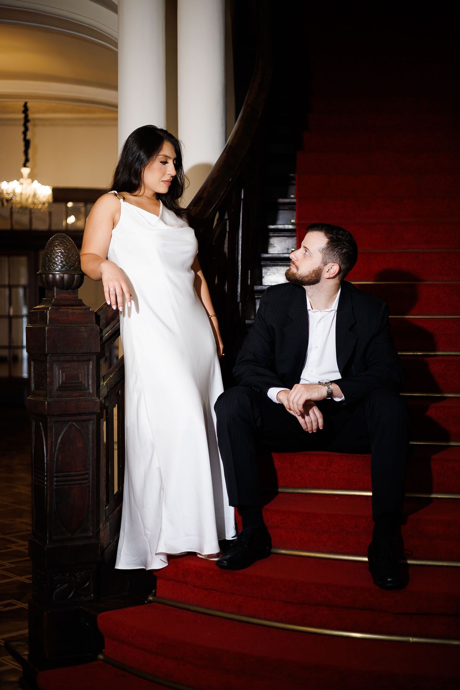 Elegant couple on red carpeted stairs, woman in white gown, man in black suit gazing up at her in grand venue.