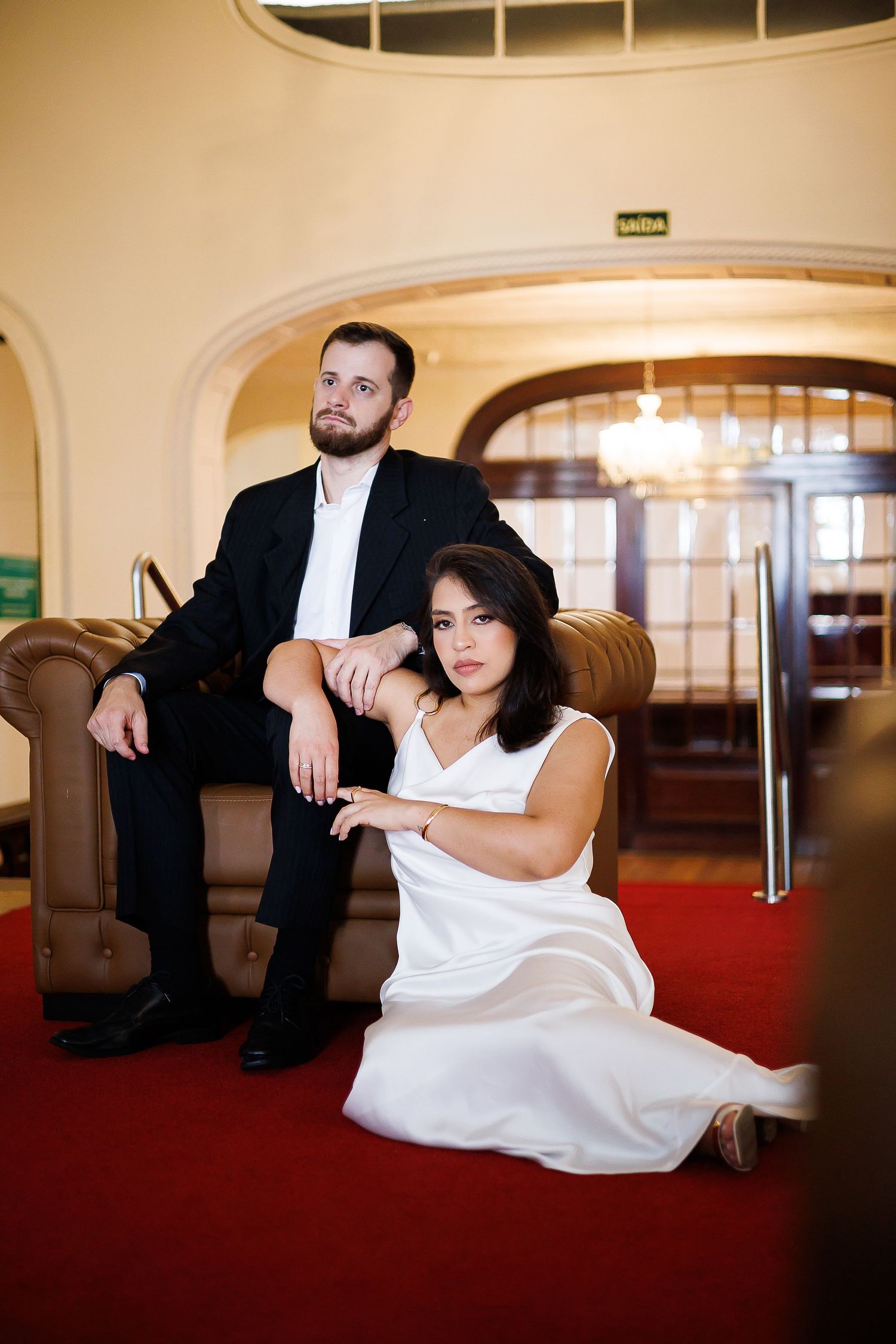 A man in a black suit sits in a leather chair while a woman in a white dress sits elegantly on a red carpet floor.