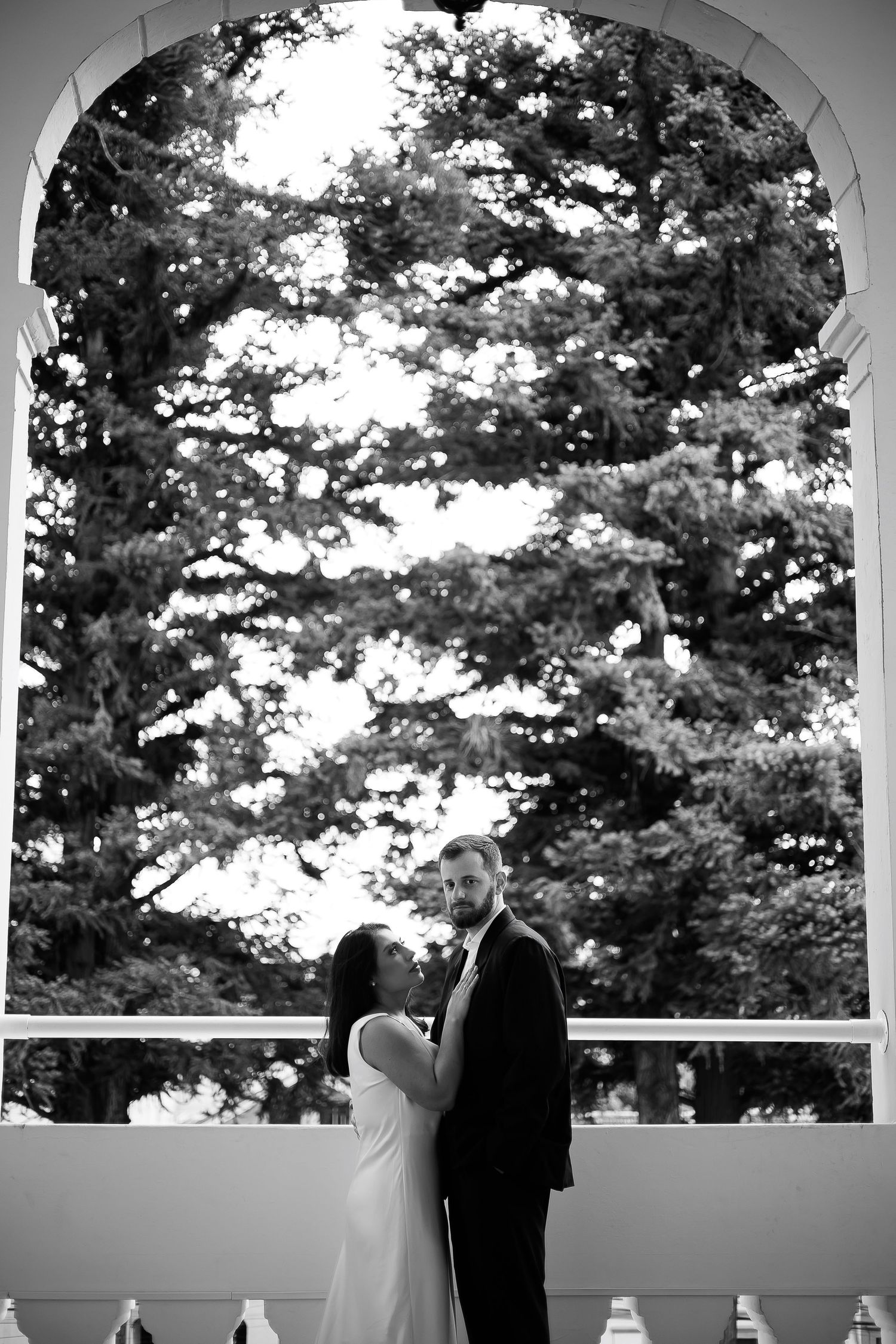 Black and white wedding photo of bride and groom embracing under an arched doorway with lush trees in the background.