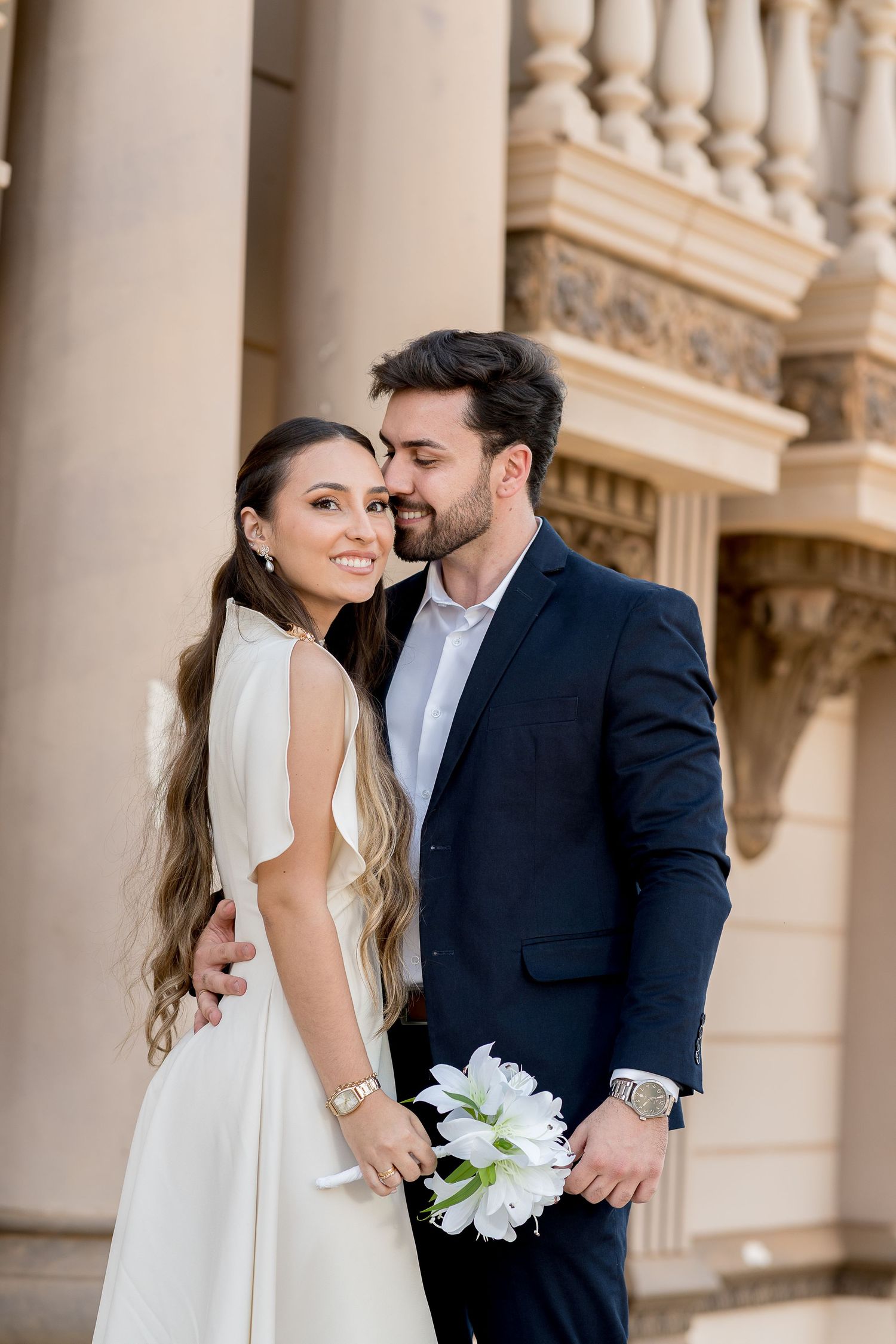 Elegant couple in wedding attire posing by classical columns, groom in navy suit kissing bride holding white flowers.