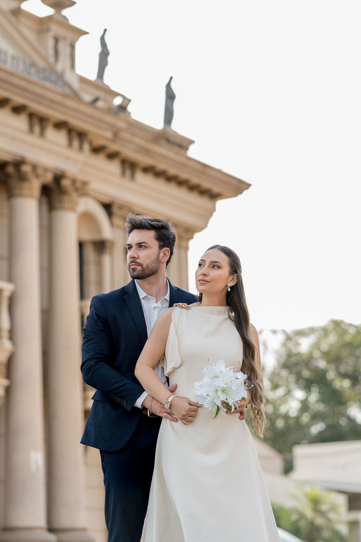Elegant bride in white gown holding bouquet with groom in dark suit posing before a classical building.