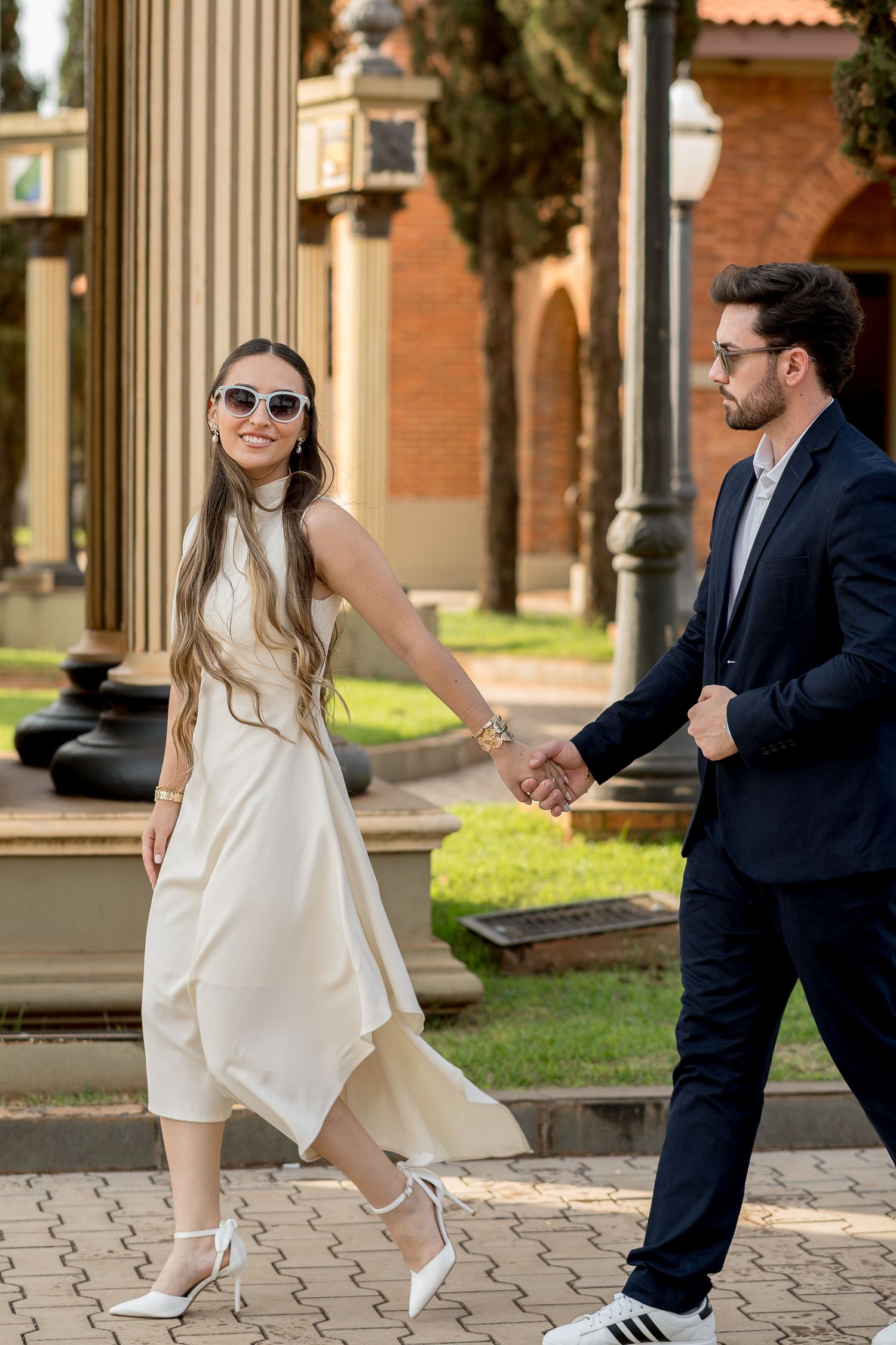 Elegant couple holding hands outdoors, woman in white dress and sunglasses, man in navy suit, walking on brick pathway.