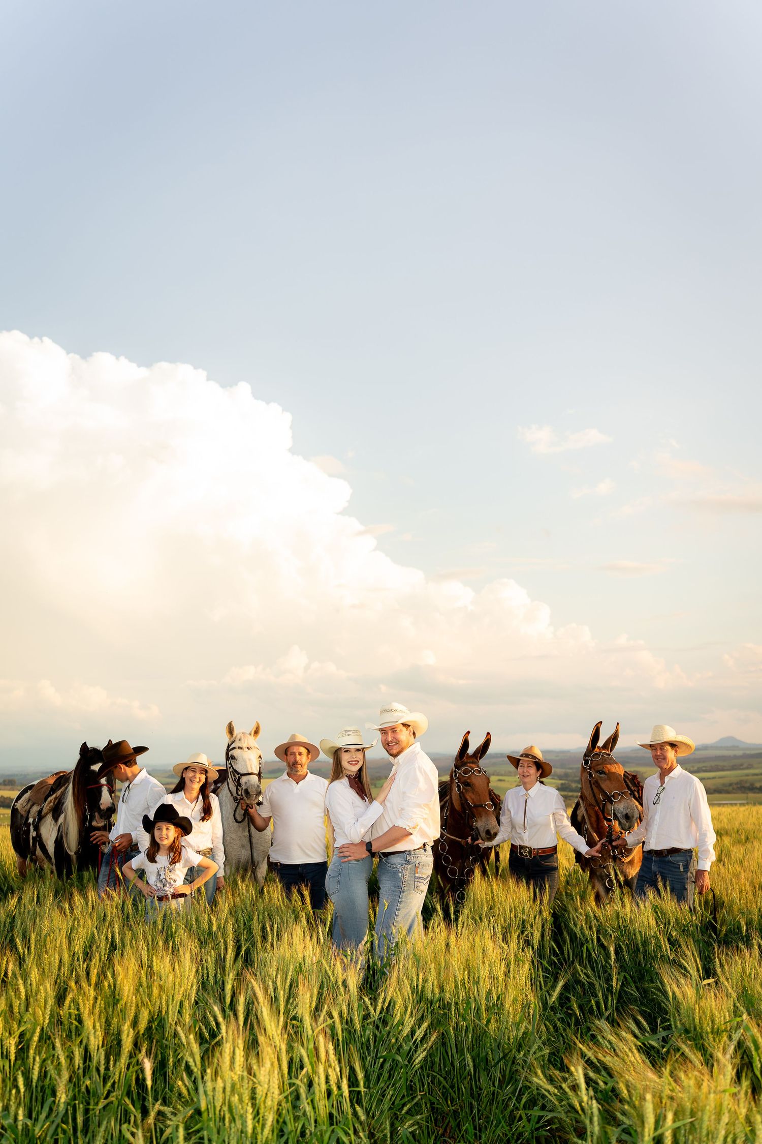 A family in white shirts and cowboy hats poses with horses in a sunlit green field under a cloudy sky.