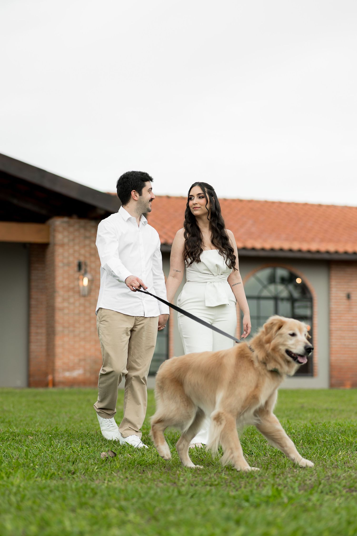 Couple in white outfits walking a golden retriever on a leash outside a brick building on green grass.
