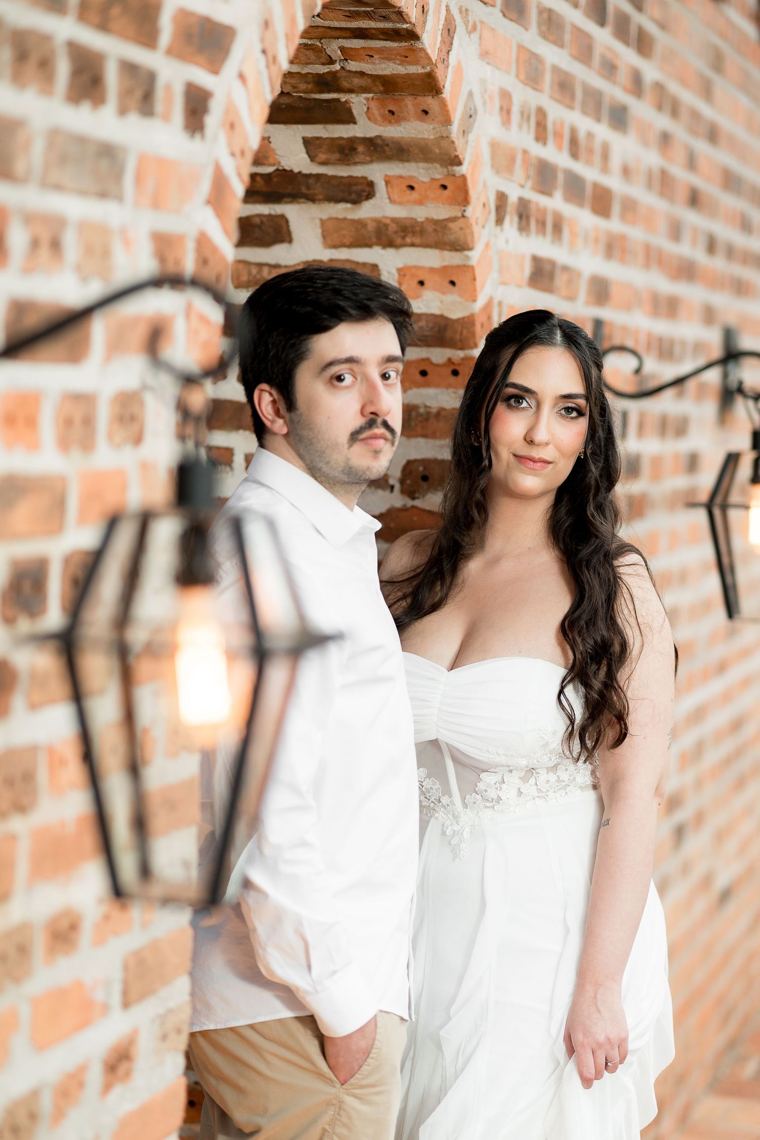 A couple in white attire poses against a rustic brick wall with vintage lanterns, evoking a romantic wedding atmosphere.