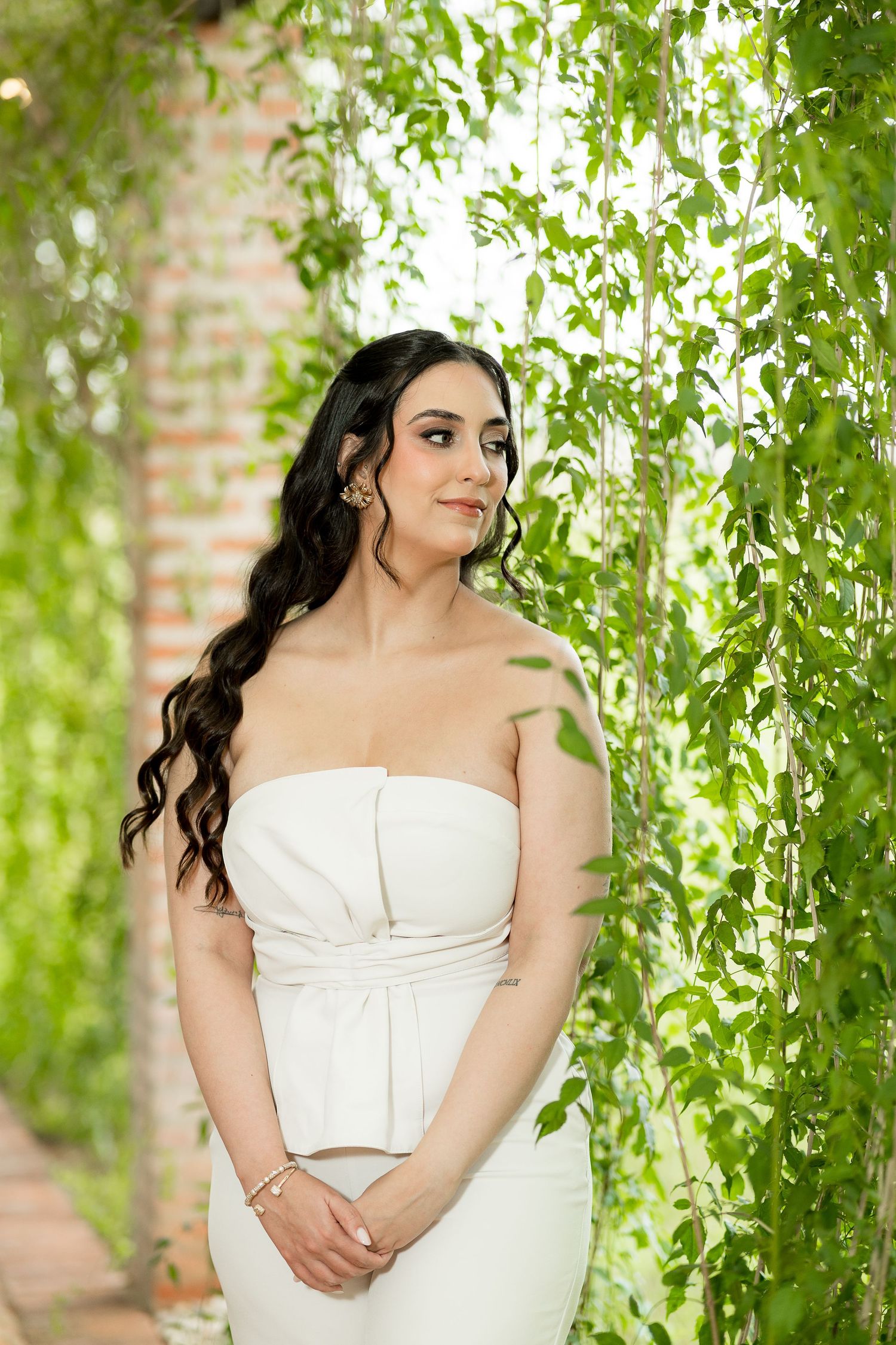 A woman in a white strapless dress poses gracefully among lush green hanging vines near a brick wall.