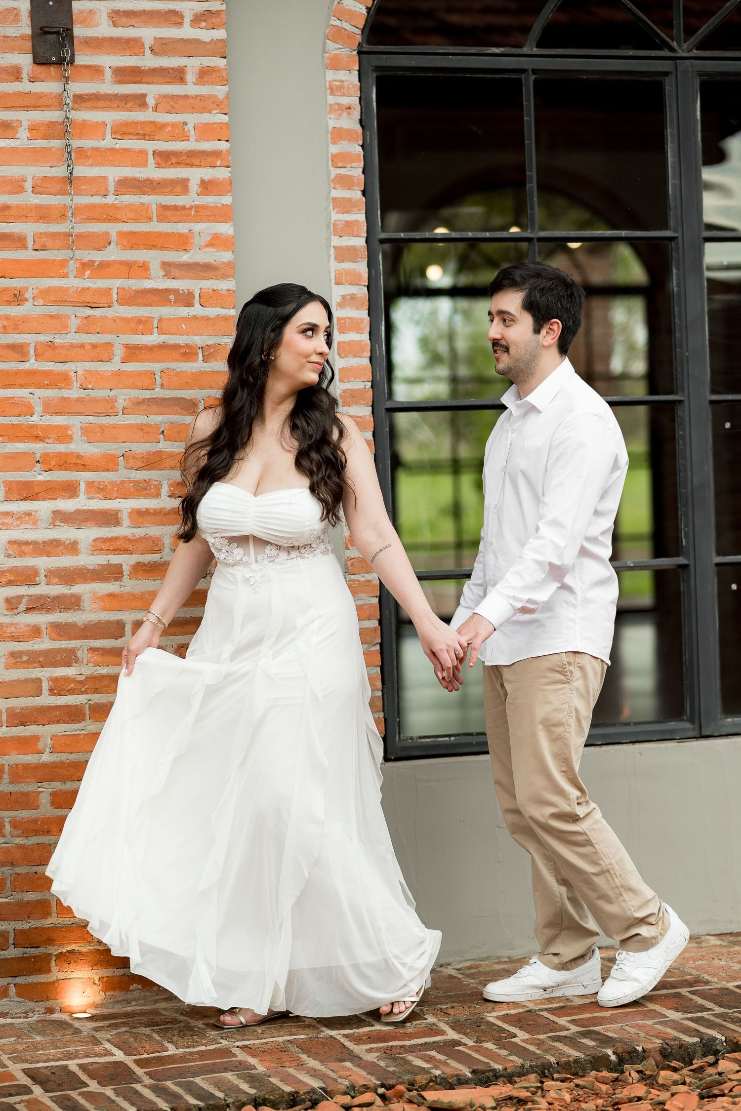 Bride in white gown and groom in white shirt hold hands, walking together beside a red brick wall.
