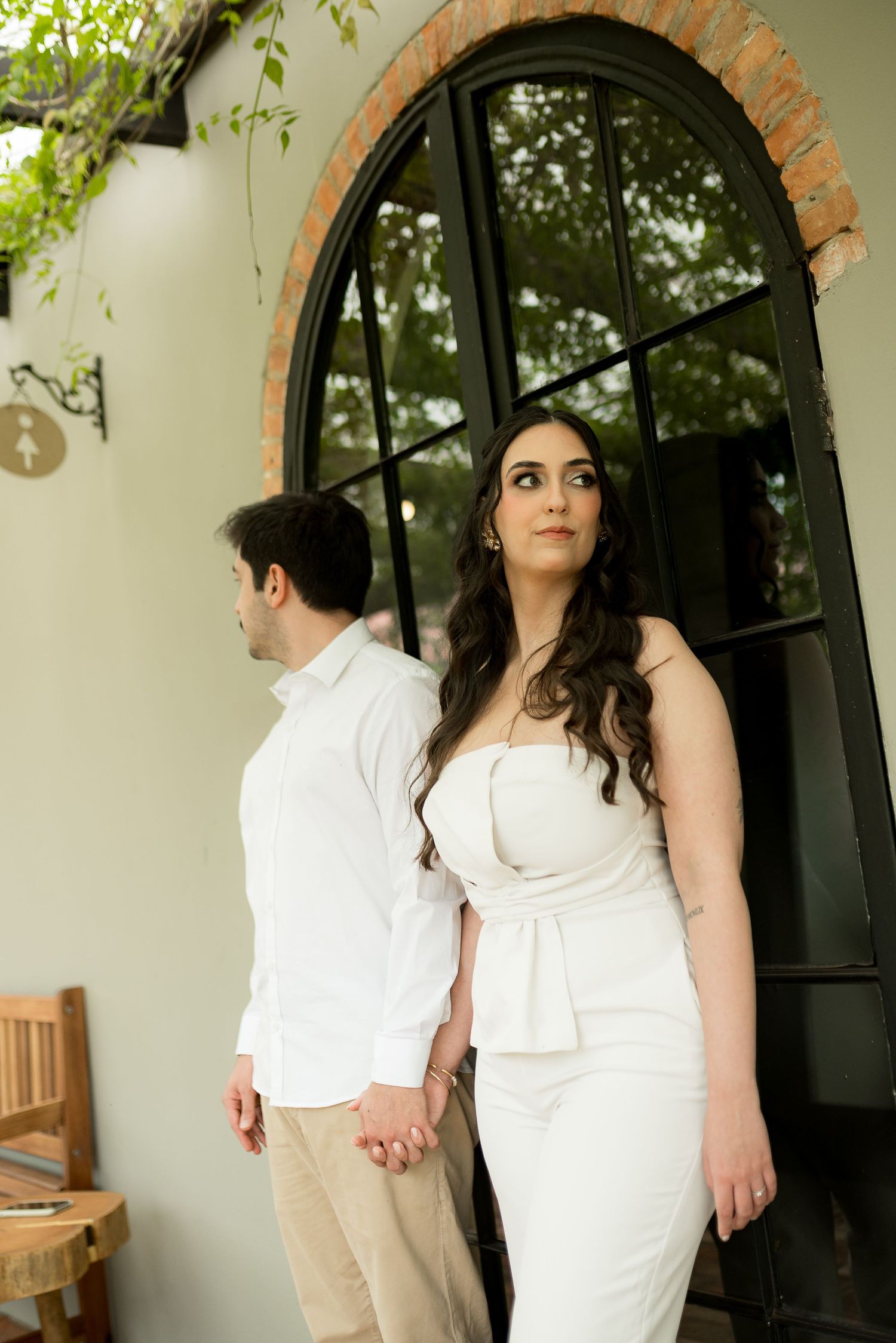 Couple in white outfits holding hands by an arched black door, woman in strapless dress gazing upward.