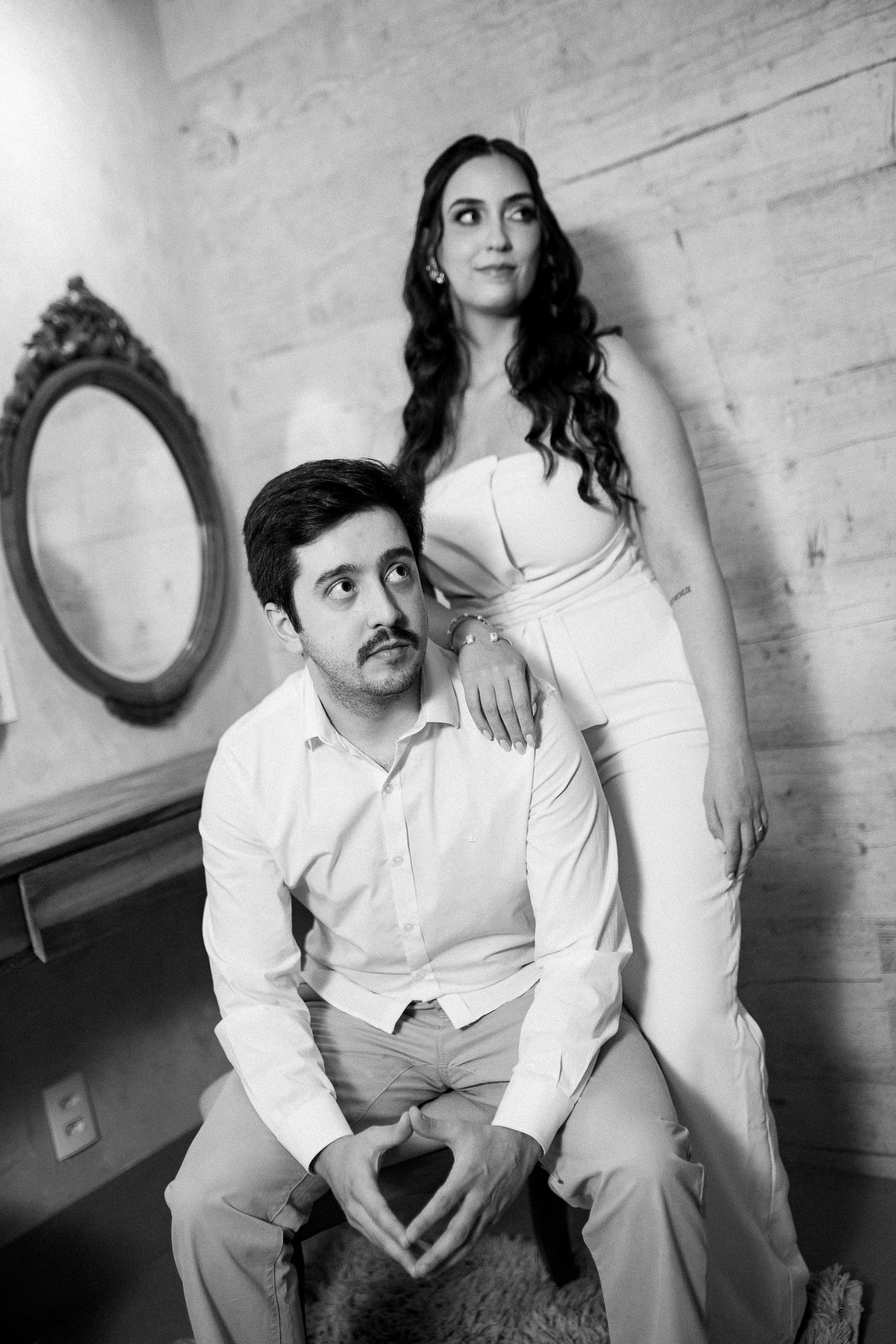 Black and white photo of a couple in white formal attire, woman standing behind seated man, elegant interior backdrop.
