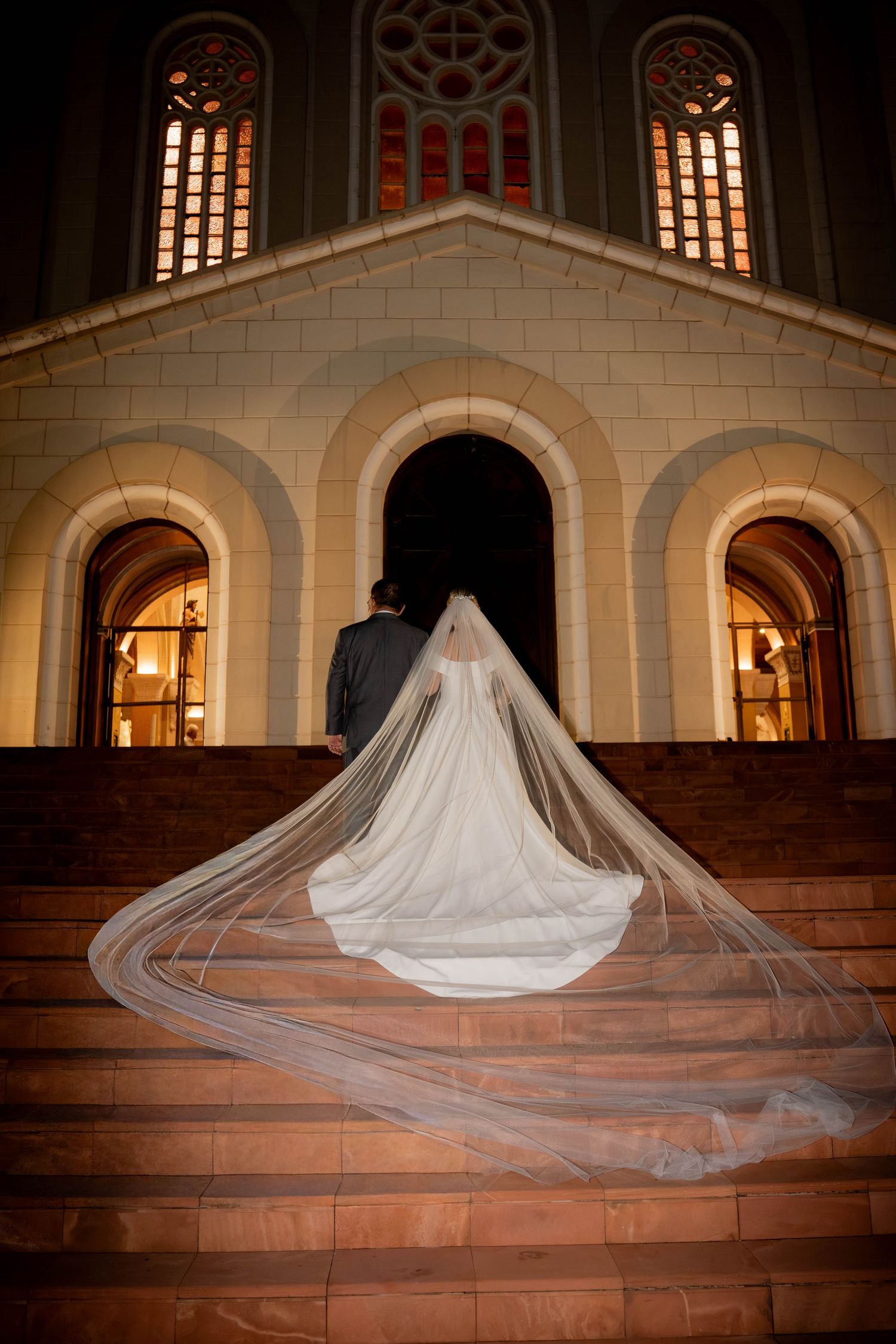 Bride and groom ascending church steps at night, her long cathedral veil dramatically spread across the stone staircase.