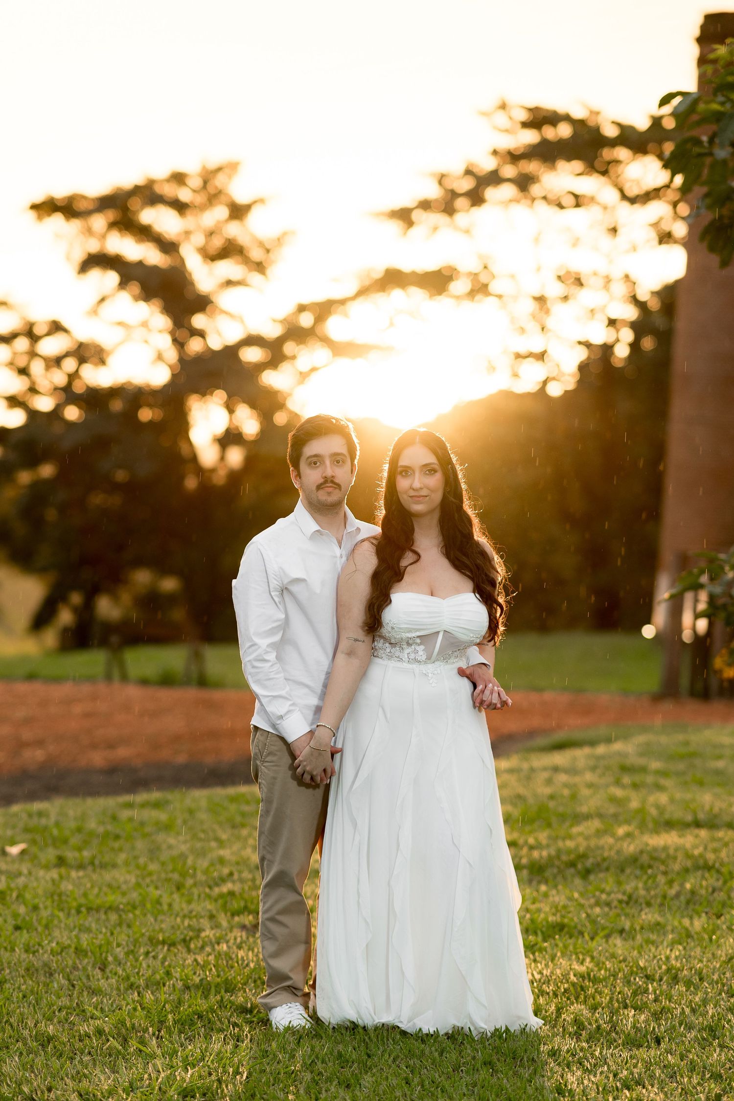 A couple in wedding attire holds hands while posing outdoors with a golden sunset glowing behind them.