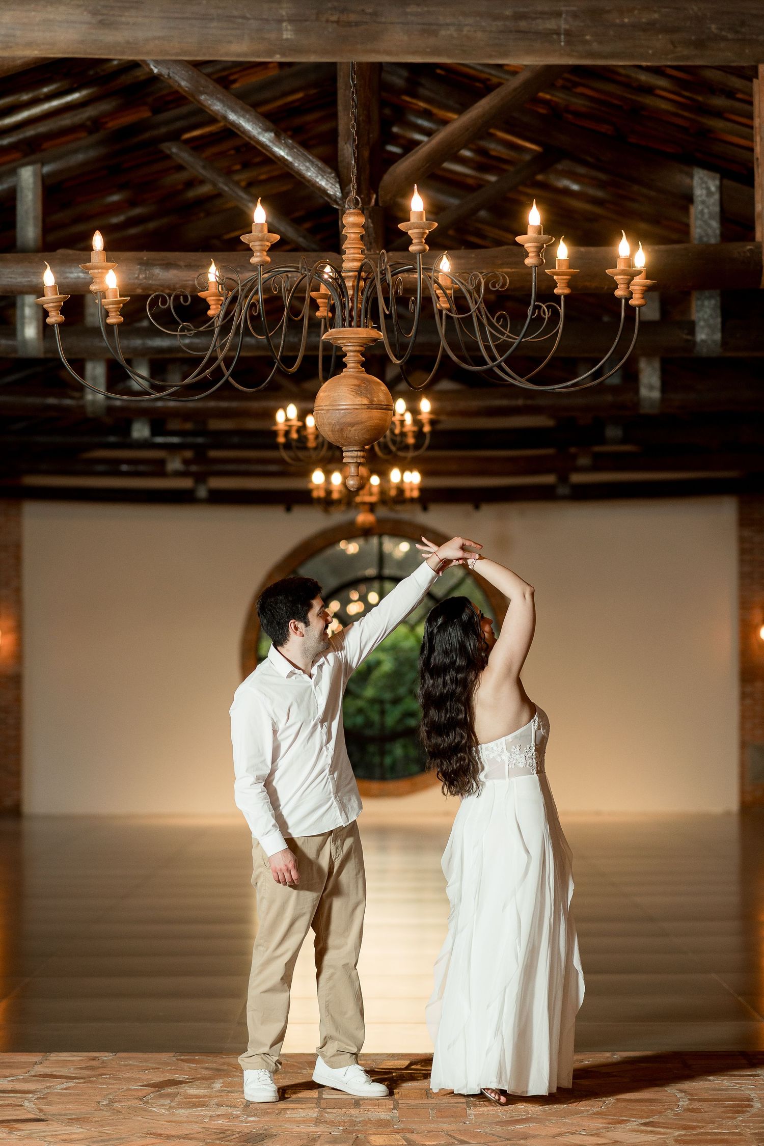 Couple dancing under chandelier in rustic barn venue, man in white shirt and woman in white dress holding hands.