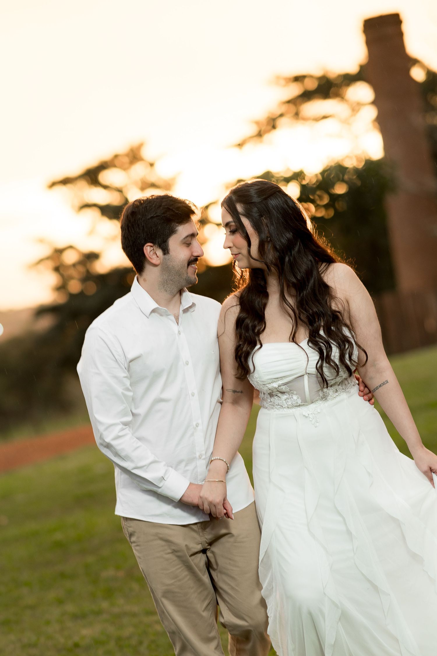 A smiling couple in wedding attire holds hands at sunset, gazing at each other on a green lawn.