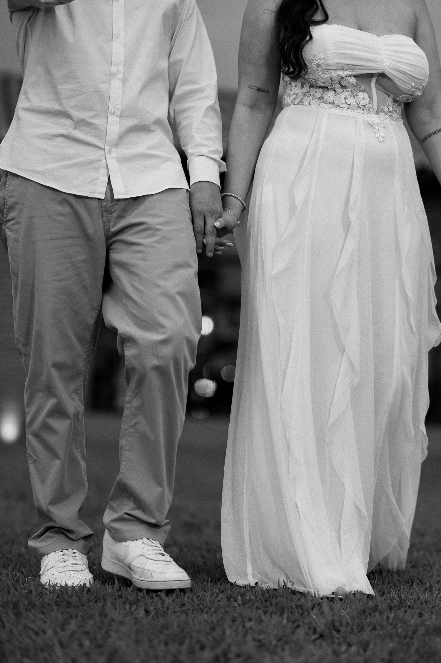 Black and white photo of a couple holding hands, bride in white gown and groom in casual attire standing on grass.