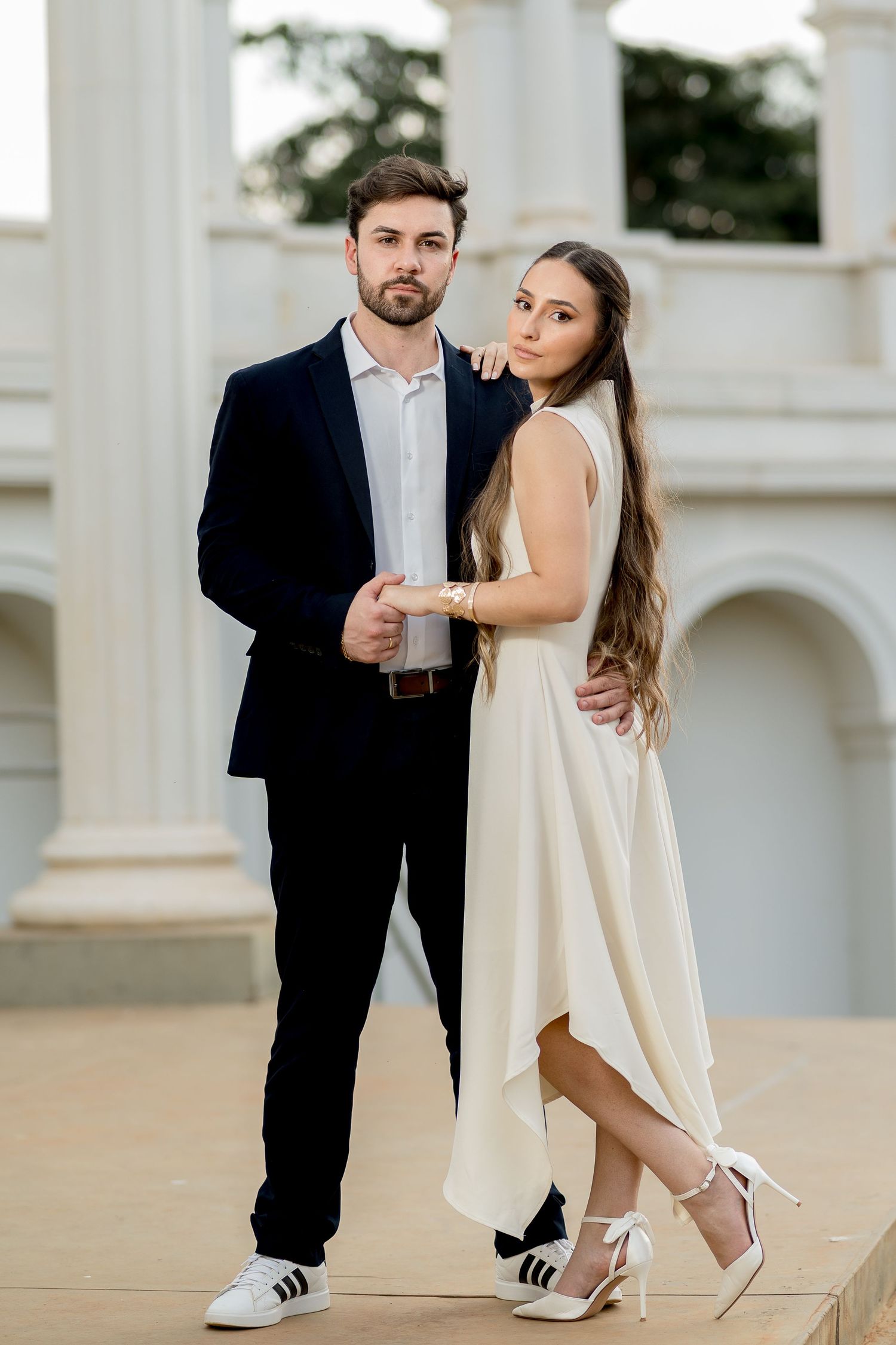 Elegant couple posing outdoors, man in dark suit, woman in white dress, near white columns.