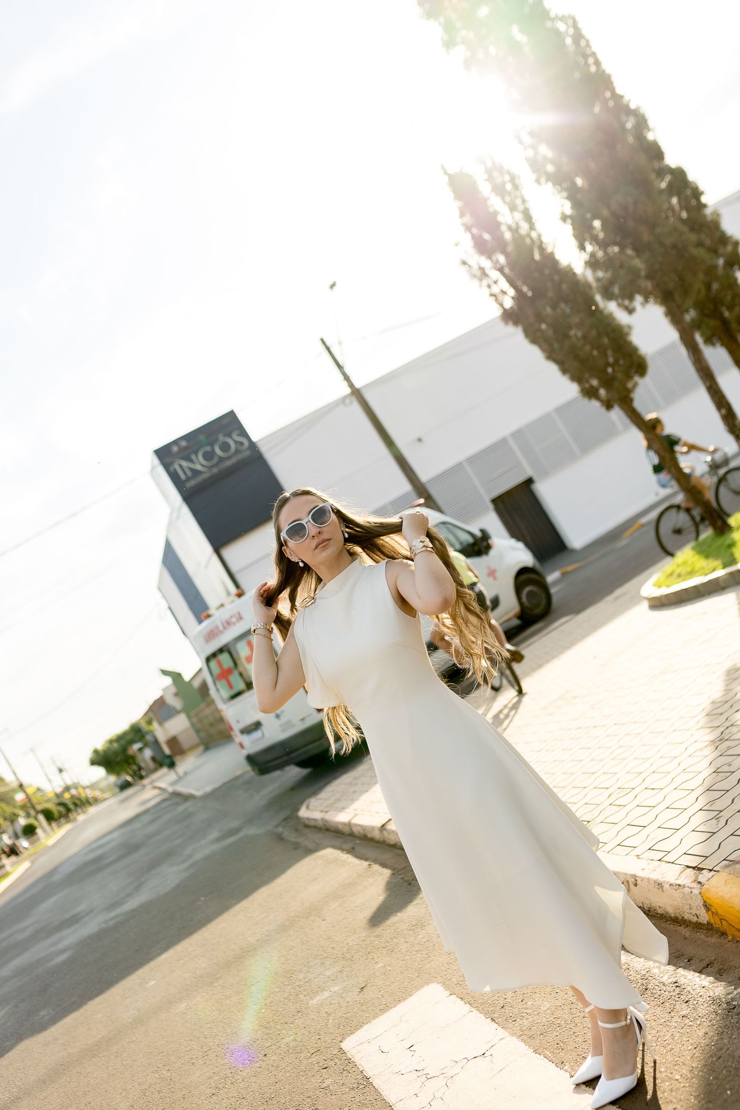 Elegant woman in white dress and sunglasses walking outdoors on a sunny day near a modern building.