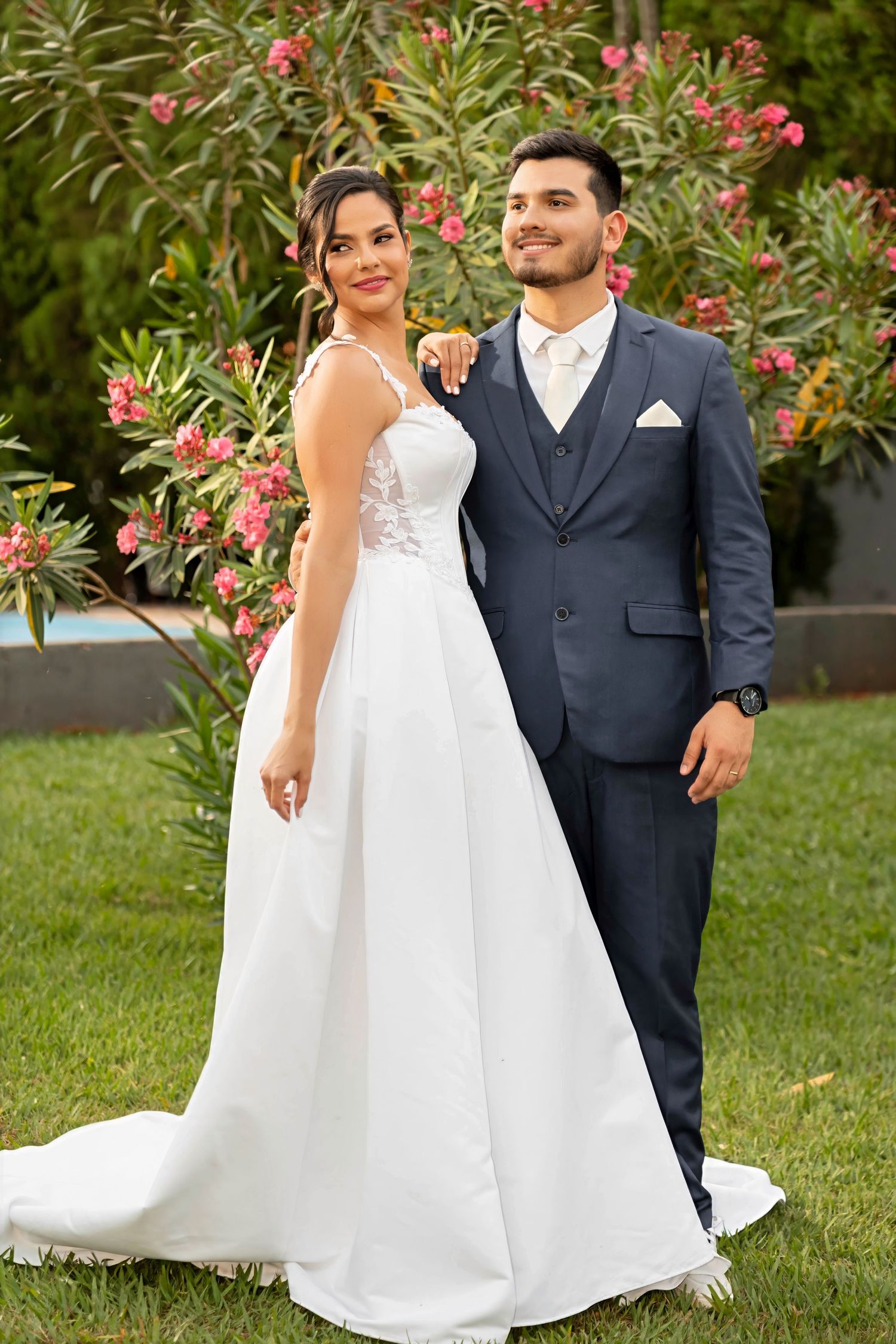 A bride in a white gown and groom in a navy suit pose together outdoors with pink flowering bushes behind them.