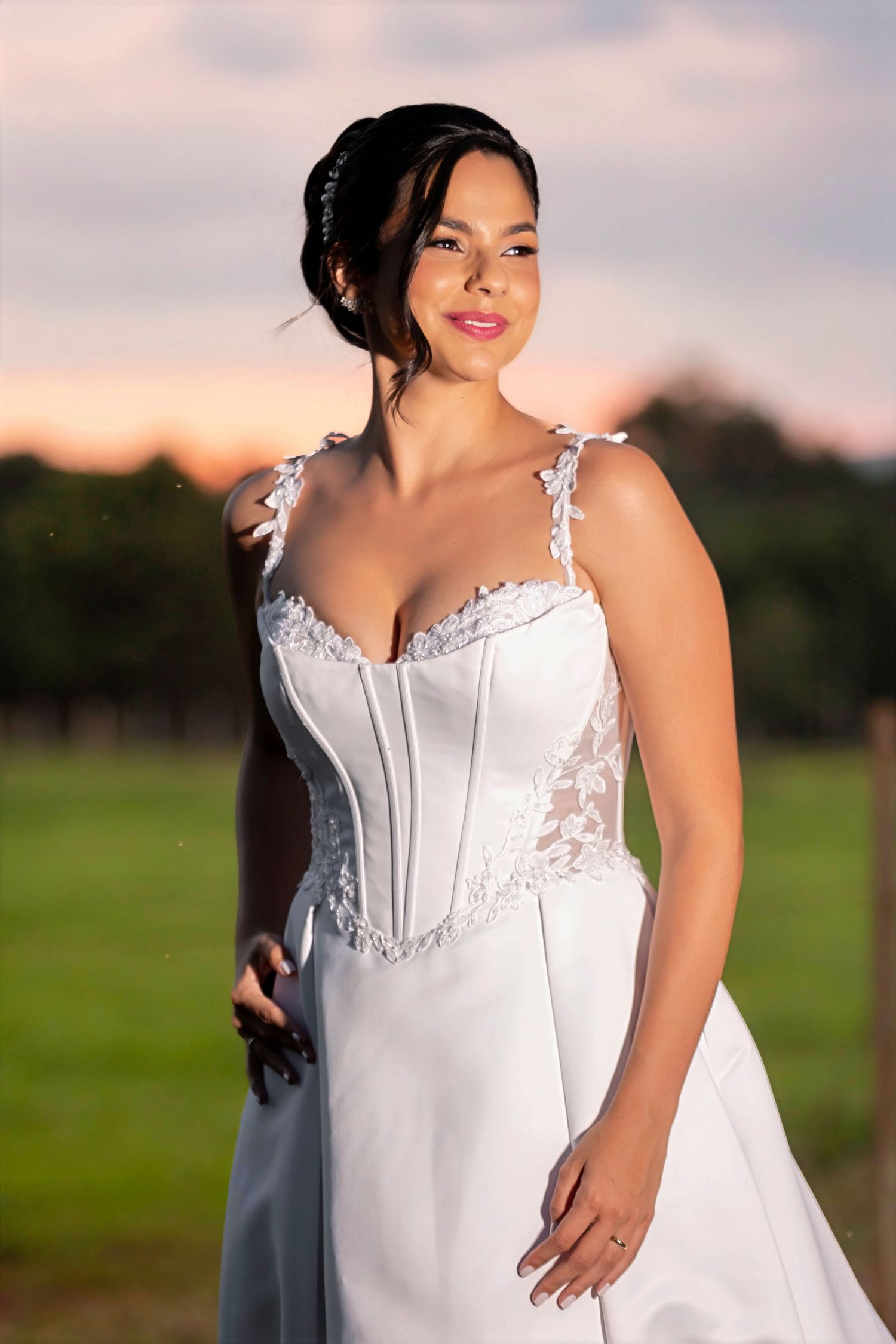 Bride in a white lace corset wedding gown poses outdoors at sunset with a soft pink sky backdrop.
