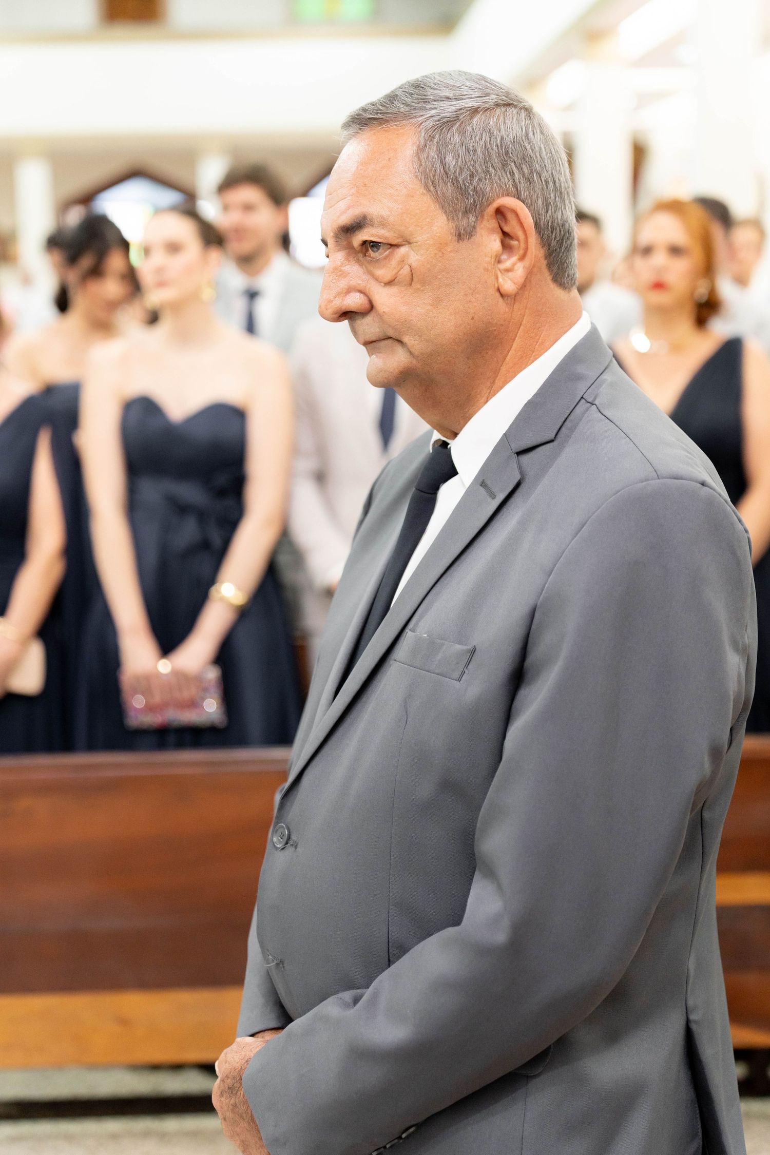Elderly man in gray suit and black tie standing solemnly in church during a formal ceremony.