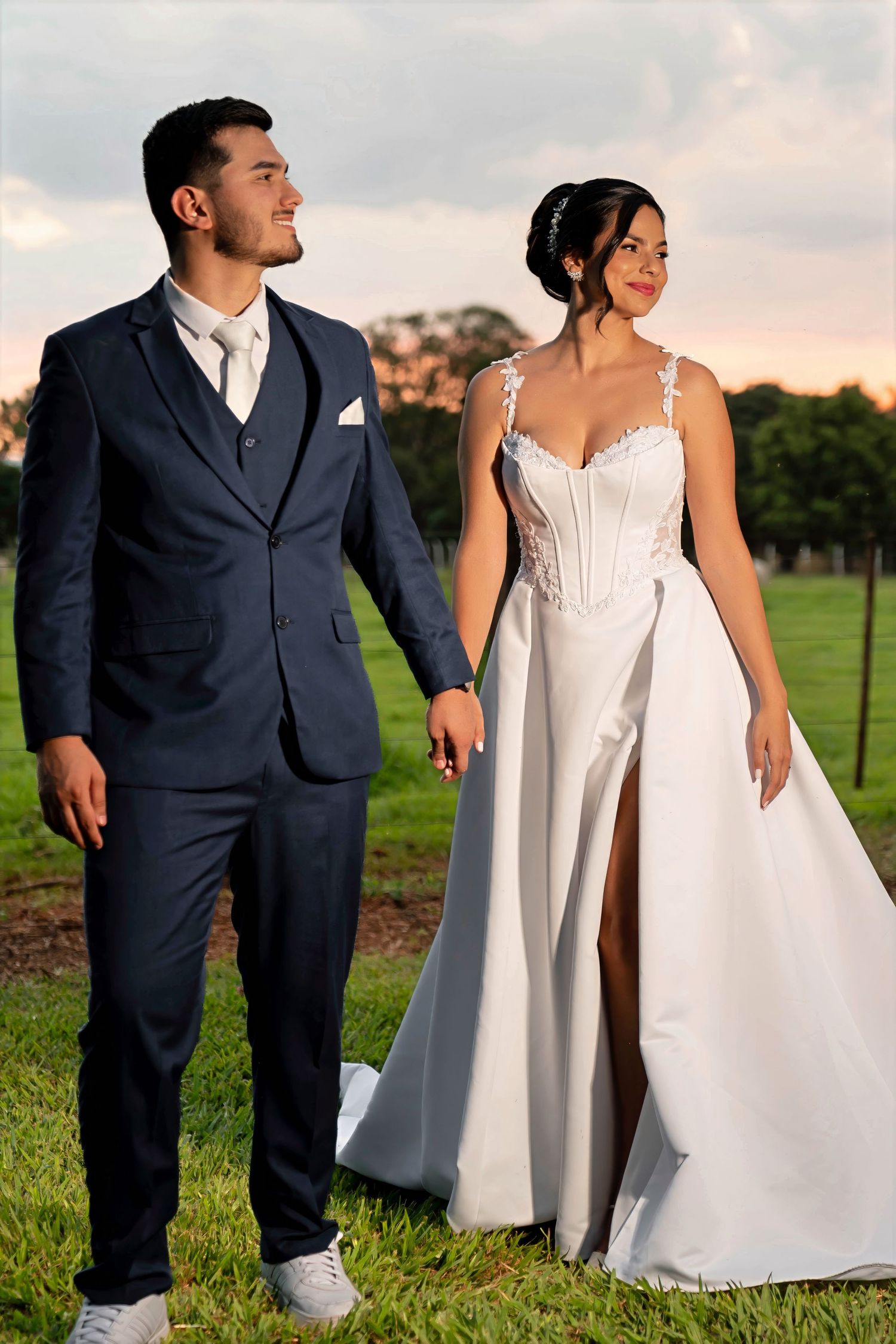 Bride in white lace gown and groom in navy suit holding hands outdoors at sunset on green grass.