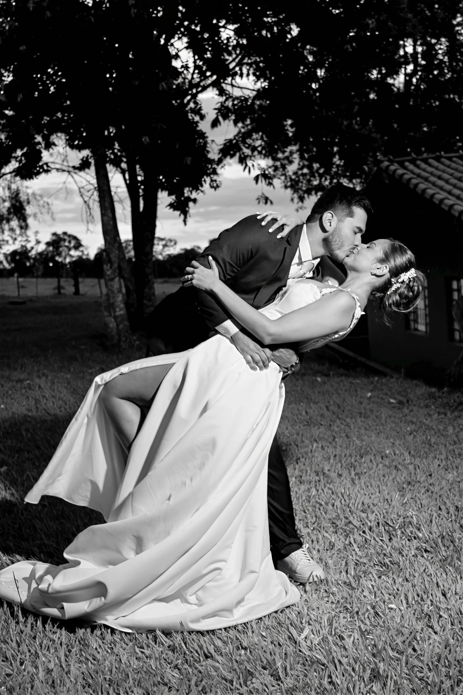 Black and white photo of a groom dipping and kissing his bride in a romantic outdoor wedding moment.