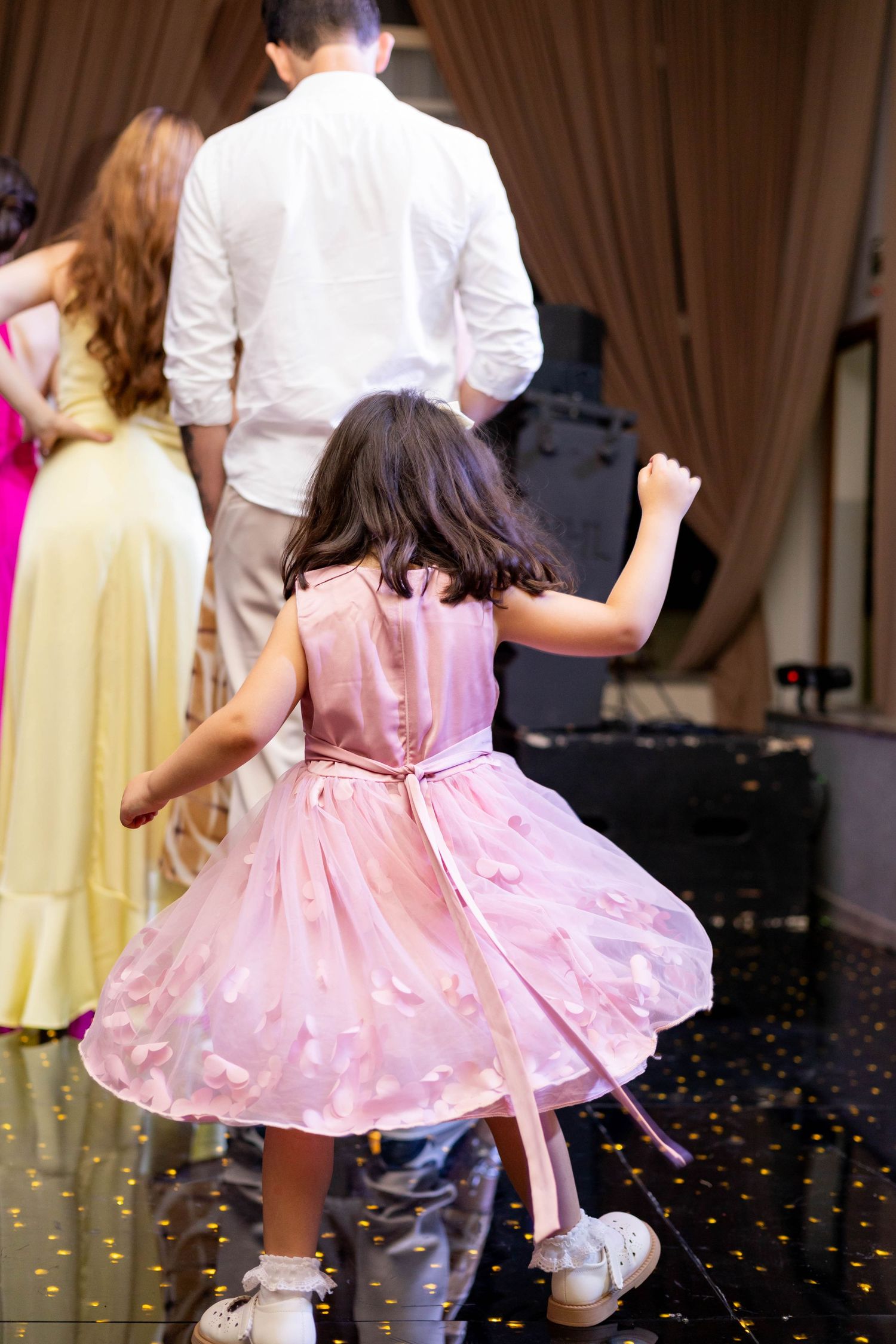 Young girl in pink dress dancing at a formal event with adults in elegant attire on a decorated dance floor.