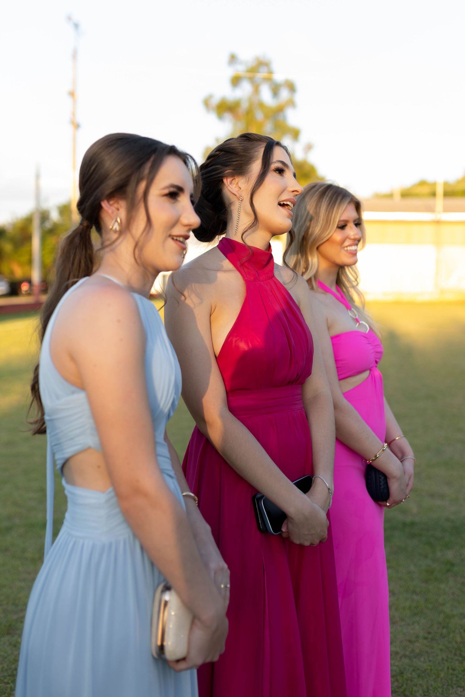 Three young women in elegant formal gowns posing outdoors at prom, wearing light blue, red, and pink dresses.