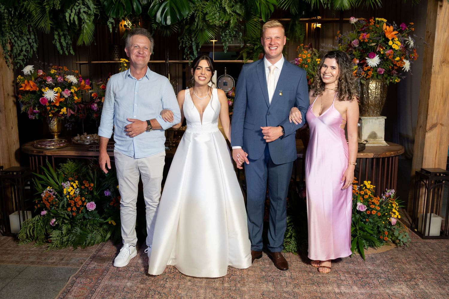 Four elegantly dressed people posing together at a floral wedding reception, including a bride in a white gown and guests in formal attire.